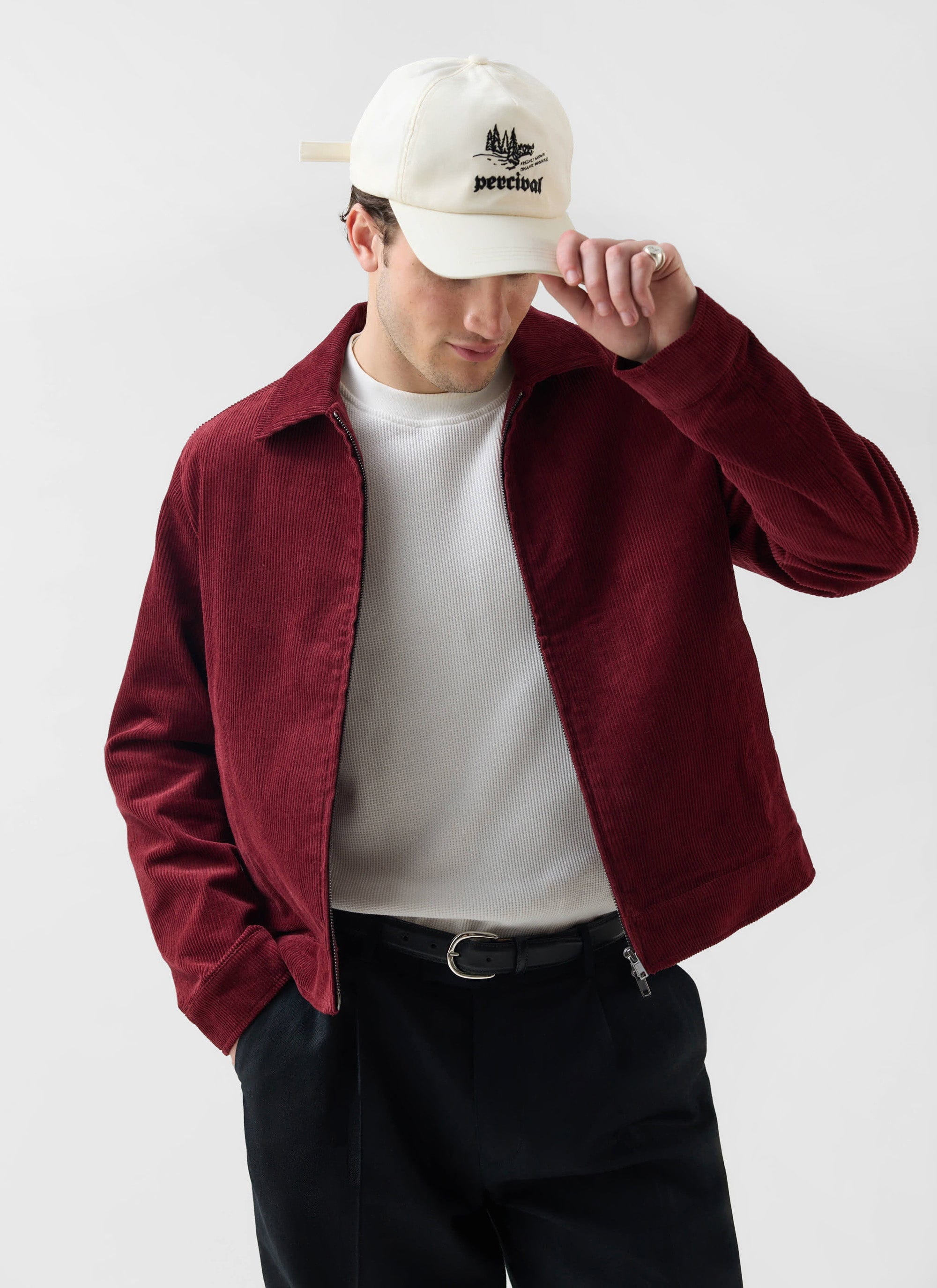 A model wears the Percival Marcel Cord Jacket in burgundy, styled with a white waffle tee and a white Percival cap.