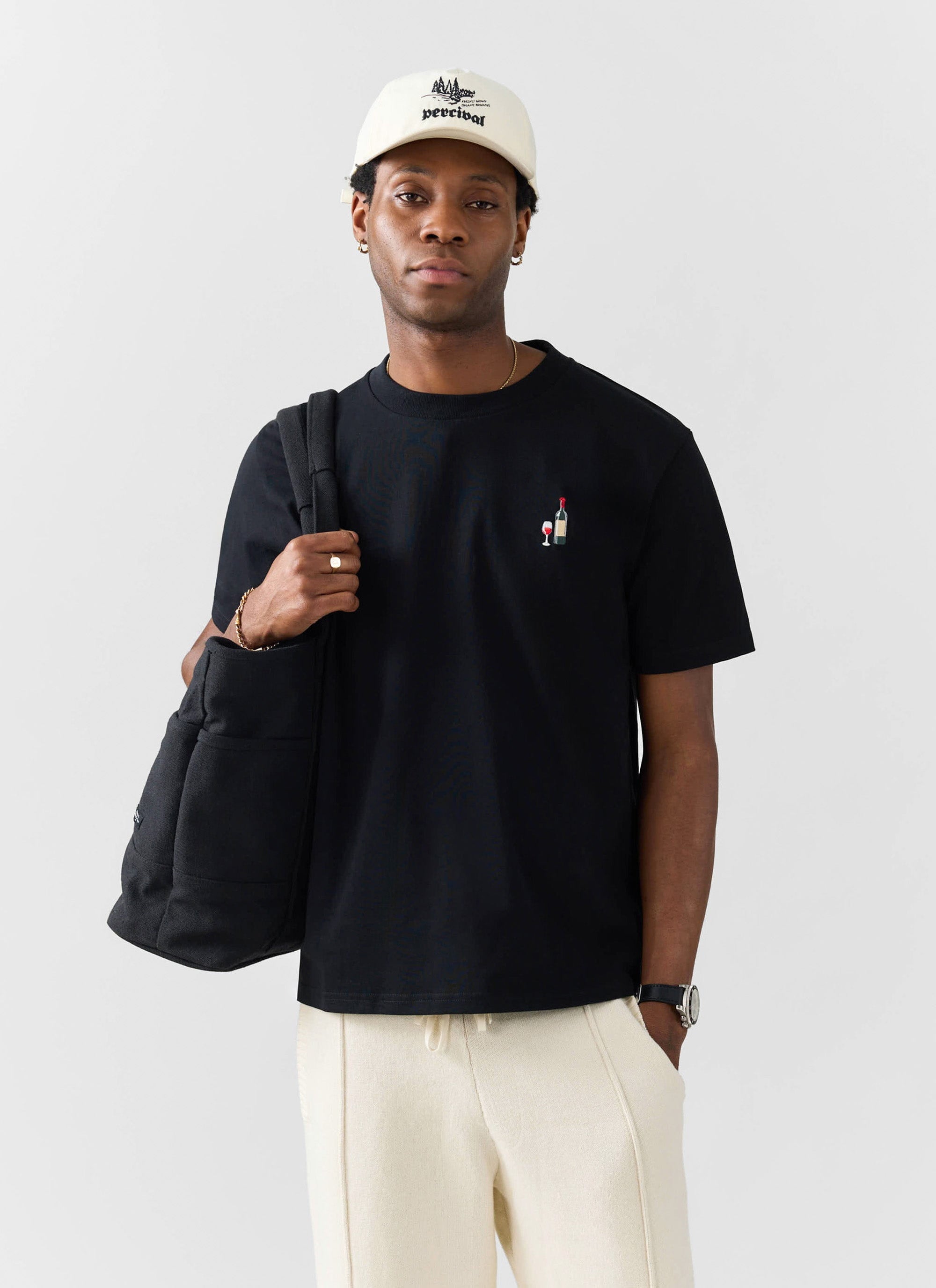 A model styles the Percival Red Wine Embroidered T-shirt in black with cream trousers and a matching Percival cap.