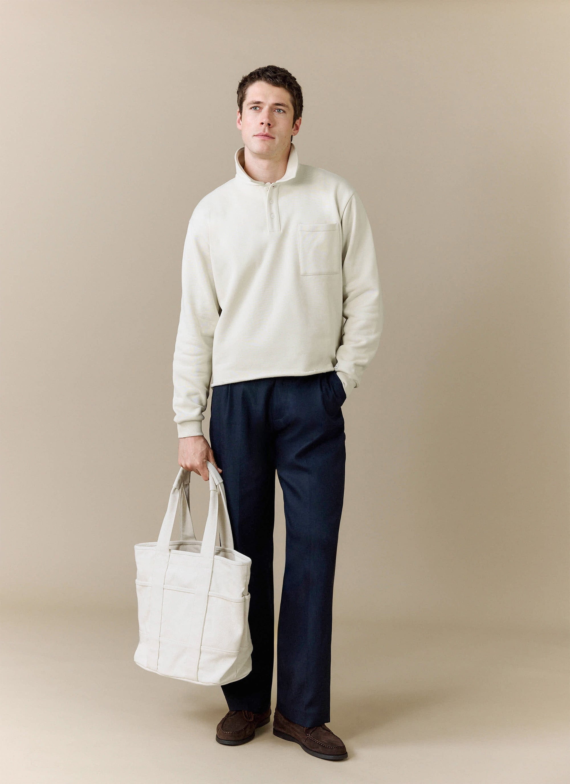 A model wears the Percival Dudley Boxy Sweatshirt in ecru, styled with navy trousers and holding a matching white tote bag.