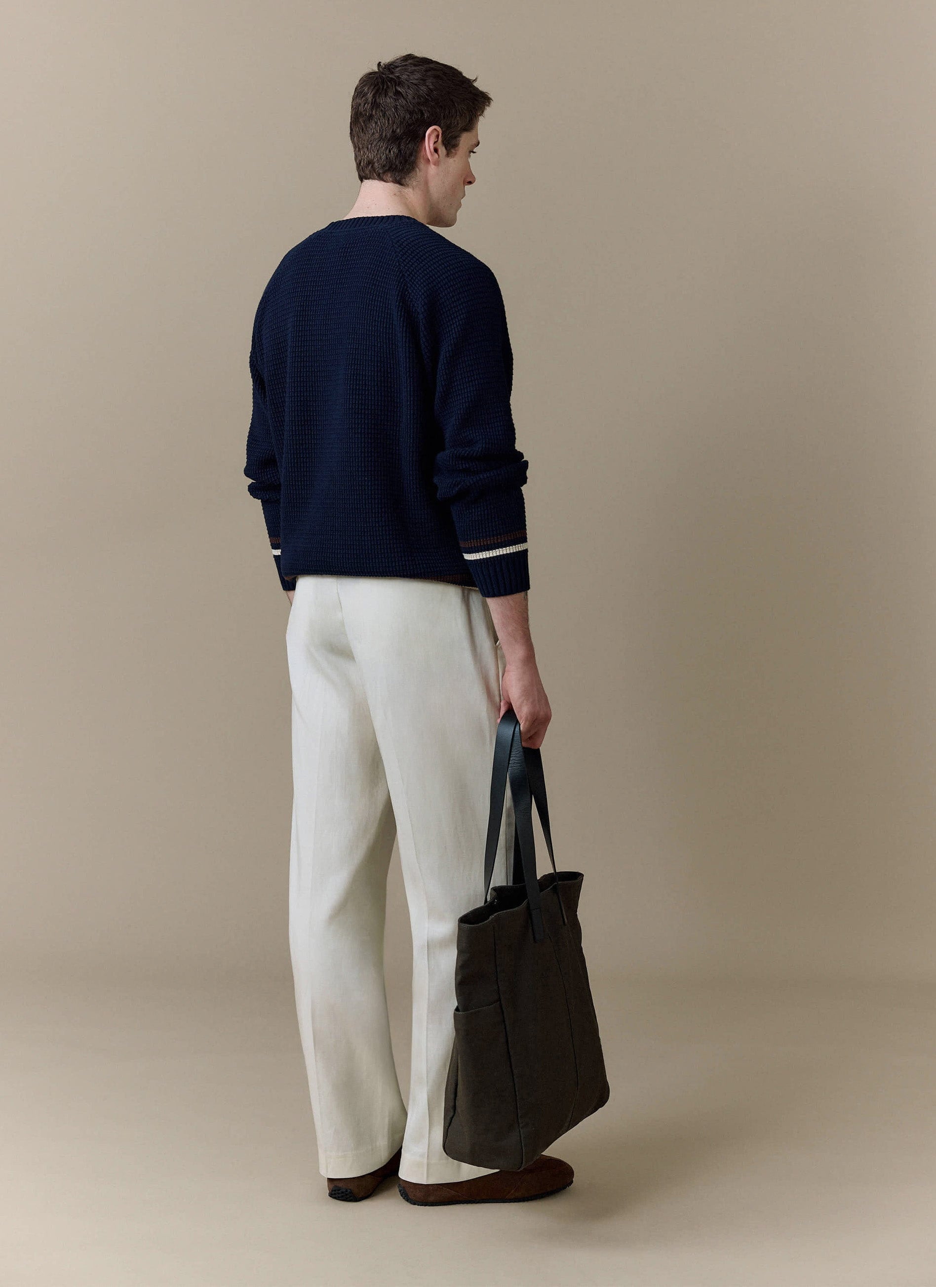 A model shot from behind wearing the navy Sergio Crochet Jumper from Percival, styled with cream trousers and a tote bag.