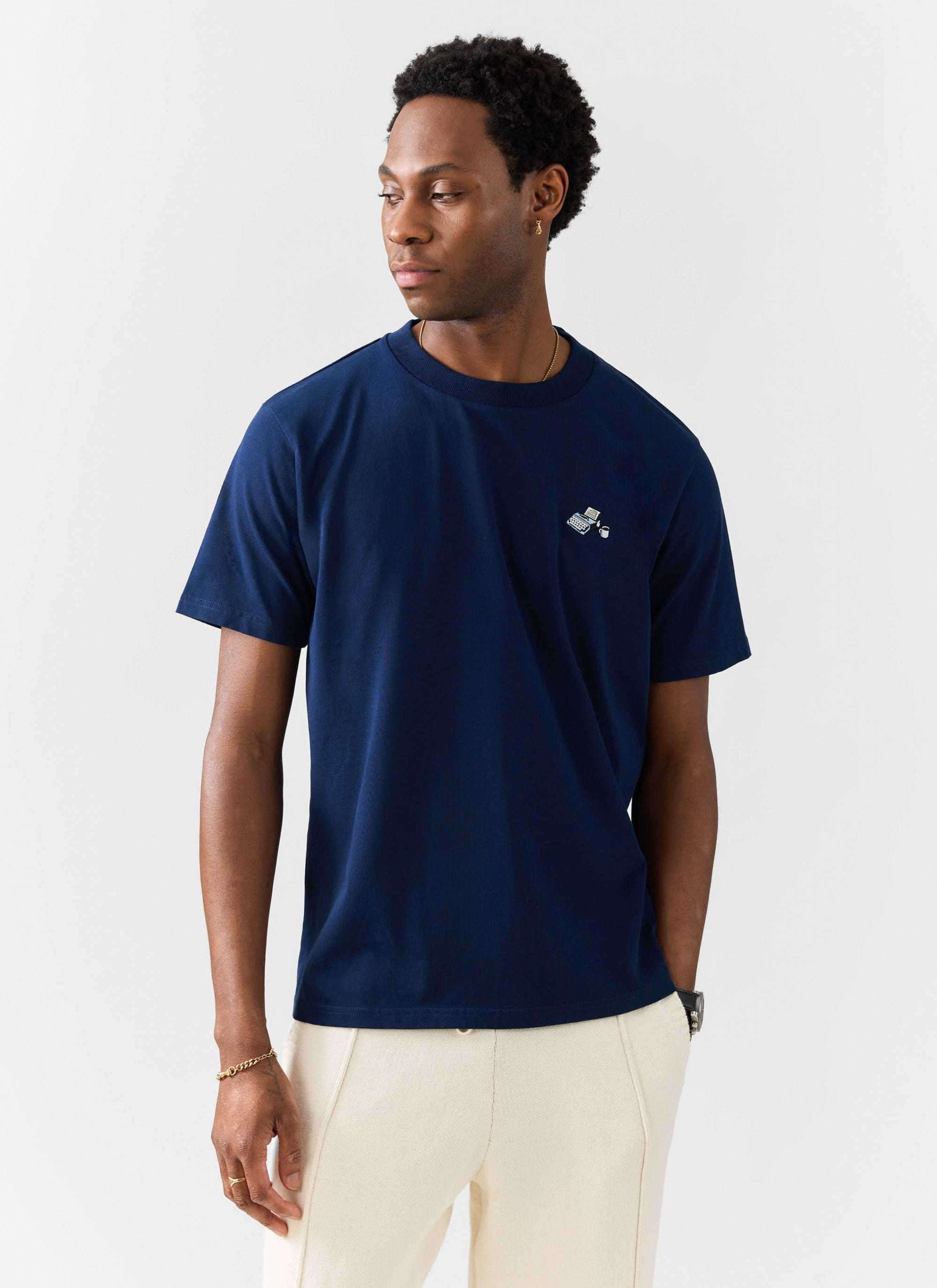 A model wears the Percival Typewriter Embroidered T Shirt in navy with cream trousers against a clean white background.