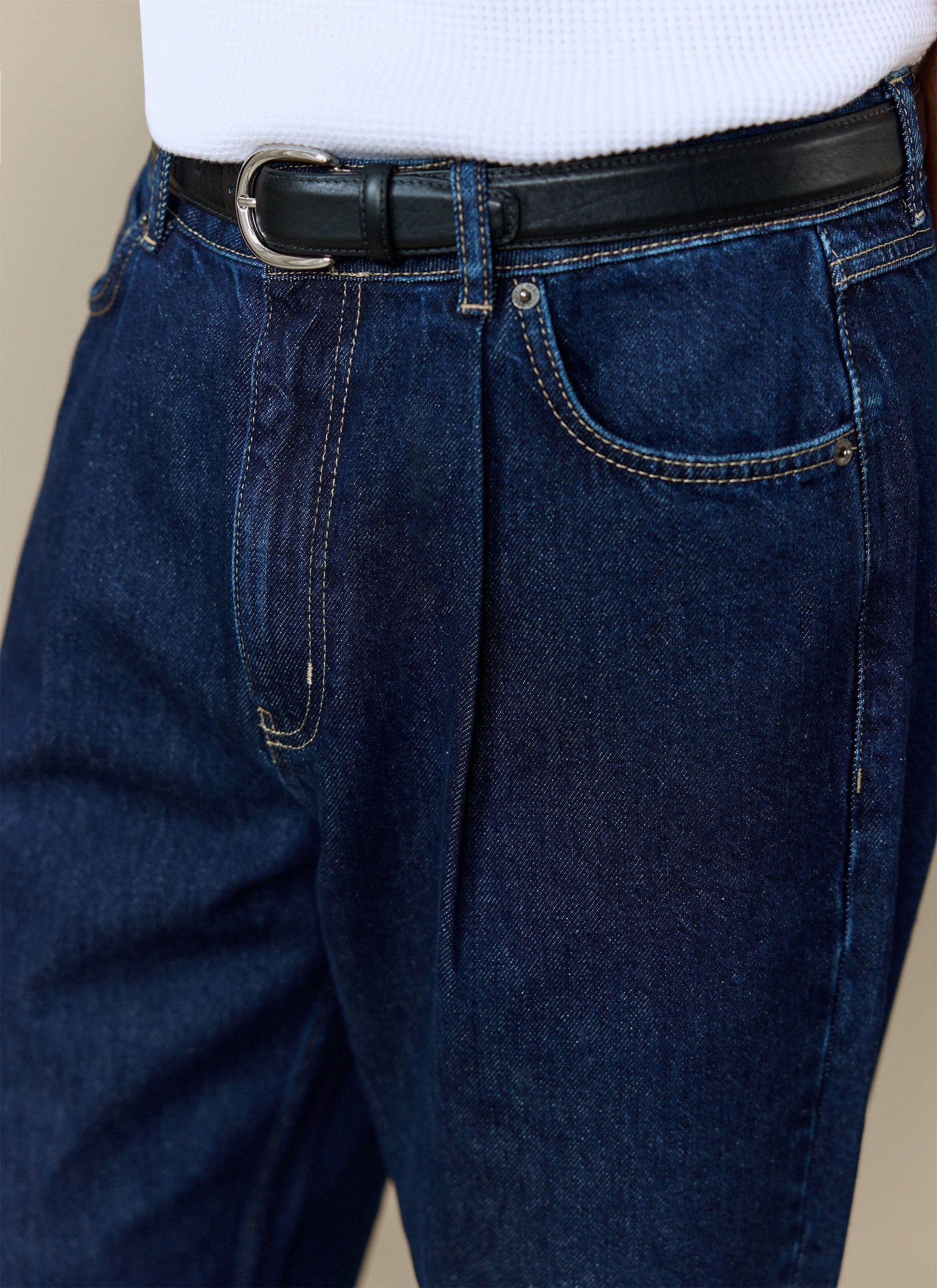 A close-up of a model wearing the Percival Pleated Denim Jeans in dark indigo, showing the front pleat and belt detail.