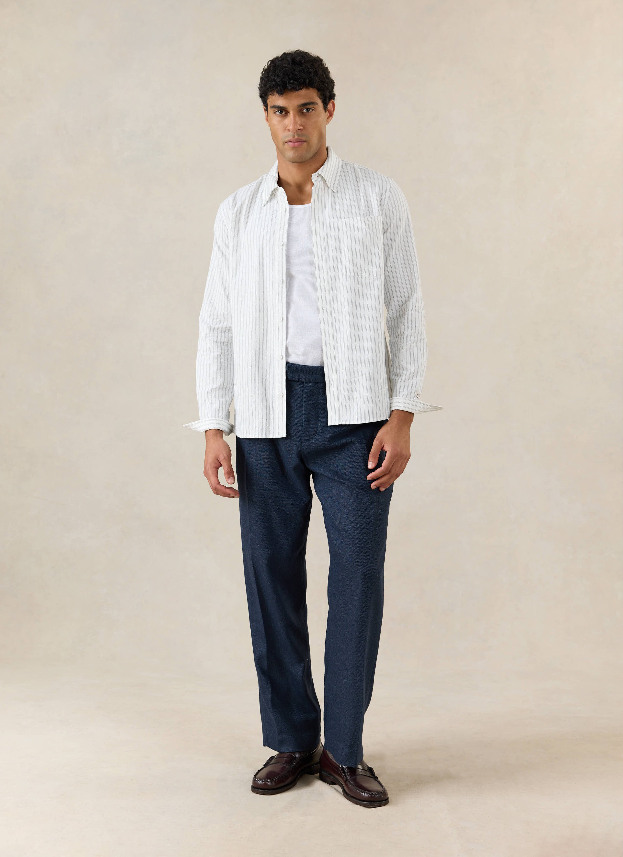 A model wears Percival Stanley Pleated Tailored Trousers in Navy Herringbone Wool, with a striped shirt and loafers.