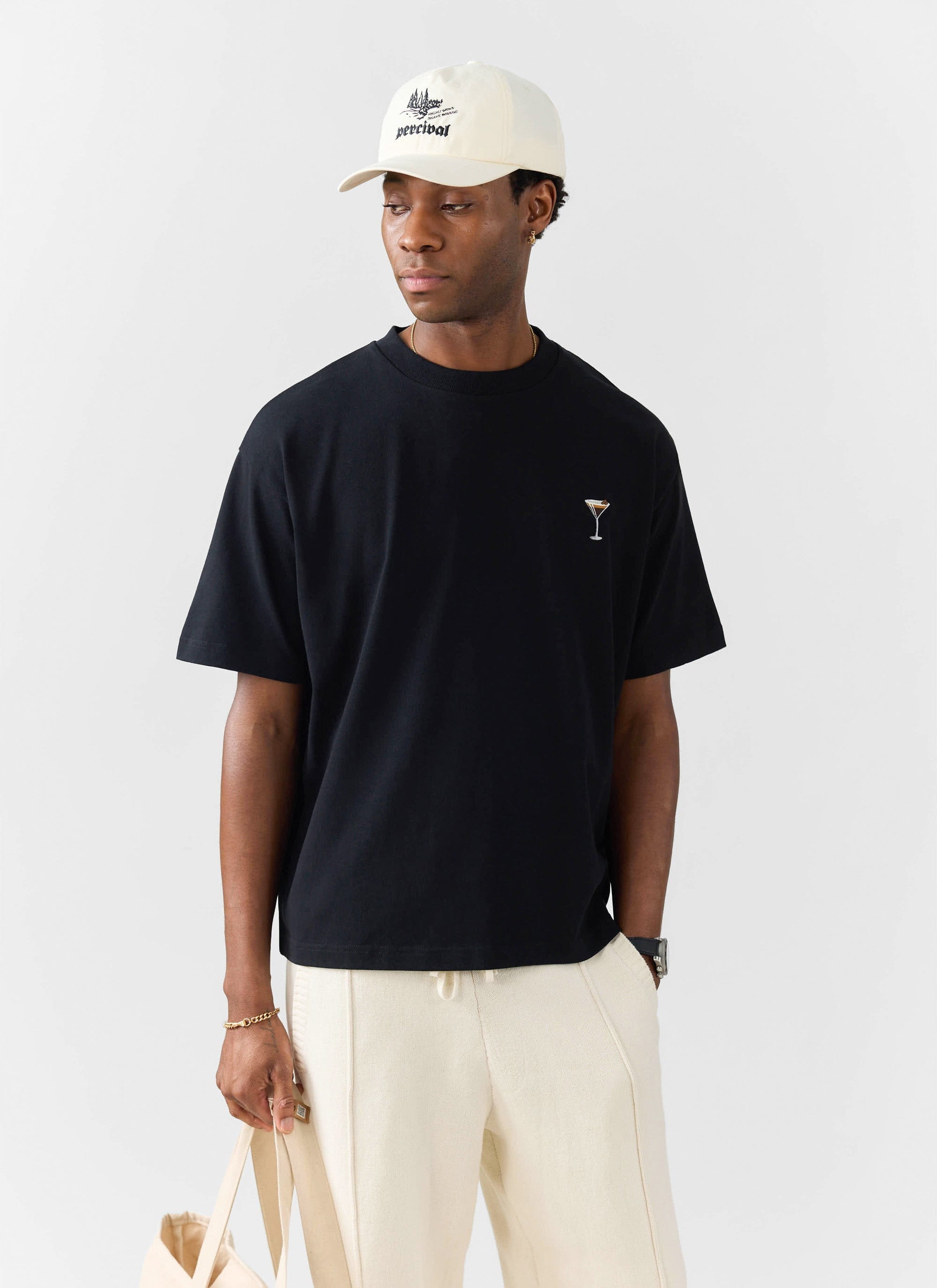A man models the black Percival Espresso Martini embroidered oversized t-shirt with cream trousers and a white baseball cap.