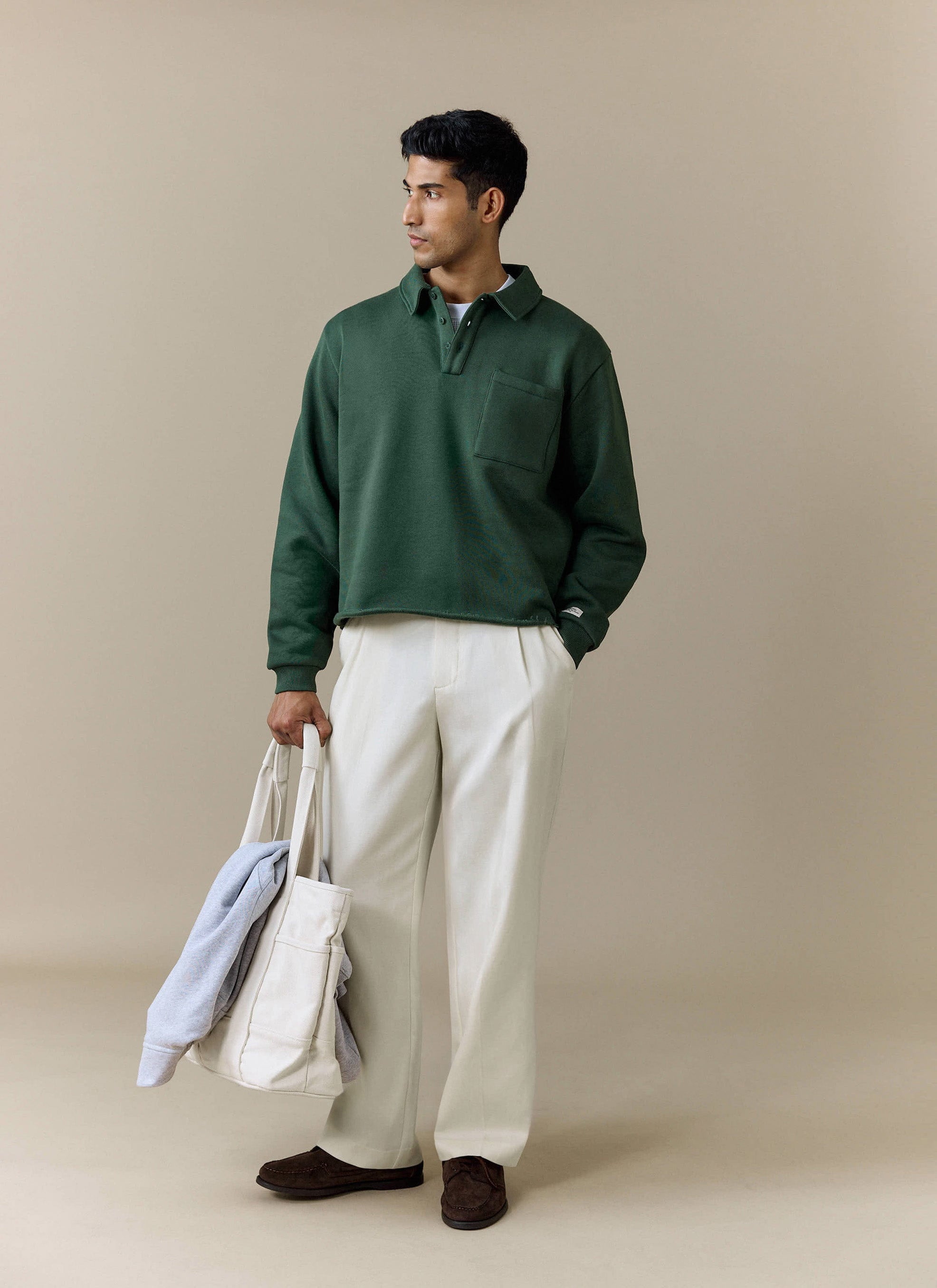 A model wears the Percival Dudley Boxy Sweatshirt in Forest, styled with white trousers and carrying a tote bag.