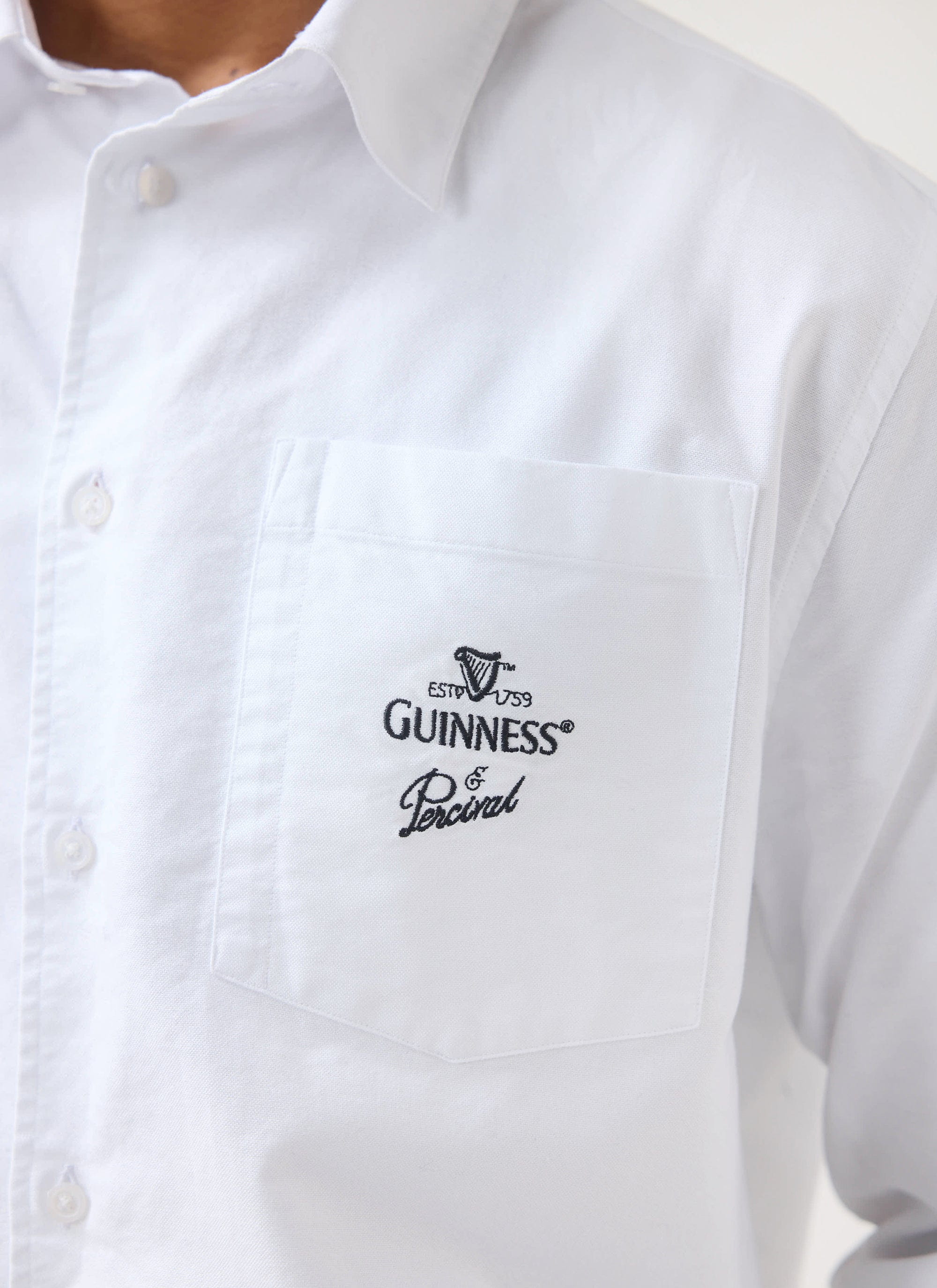 A close-up of the Guinness x Percival embroidery on the chest pocket of the white boxy button-down shirt from Percival.