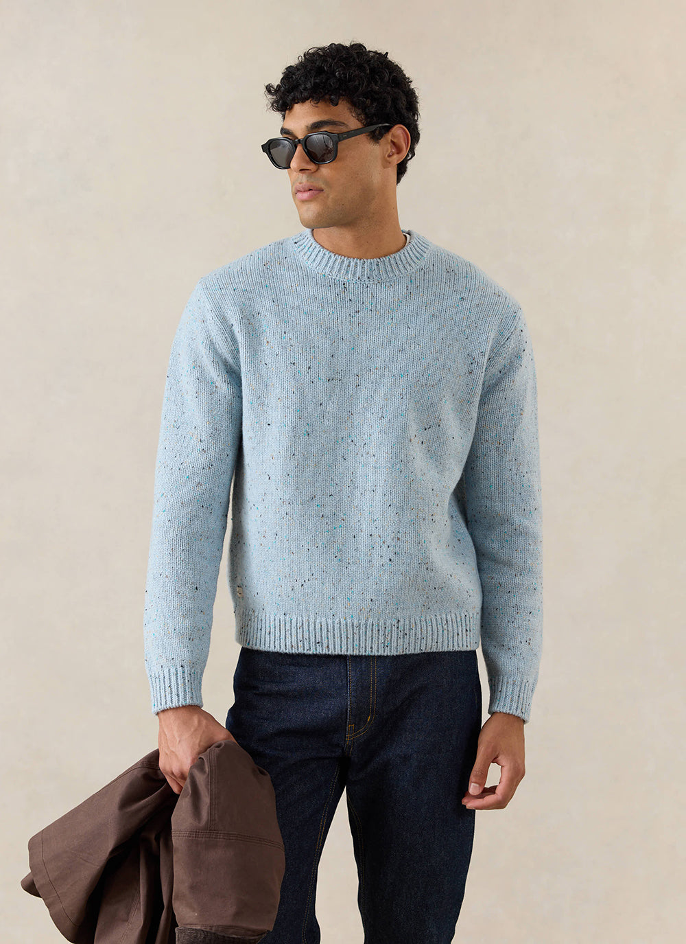 A model wears the blue Rhodes crew neck jumper from Percival, paired with dark jeans and sunglasses.