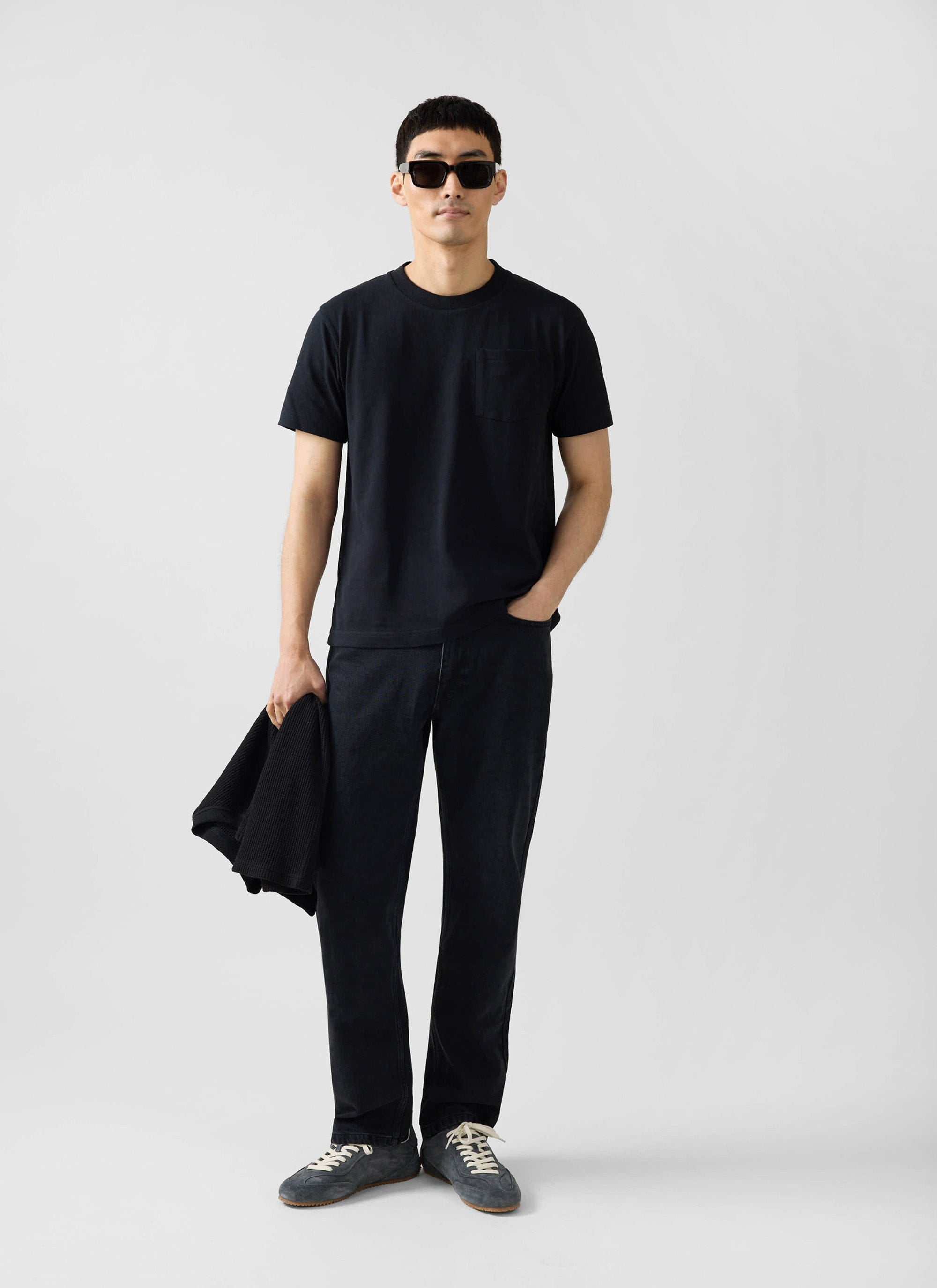 A man models the Percival Short Sleeve Pocket T Shirt in black, paired with black jeans and sunglasses.
