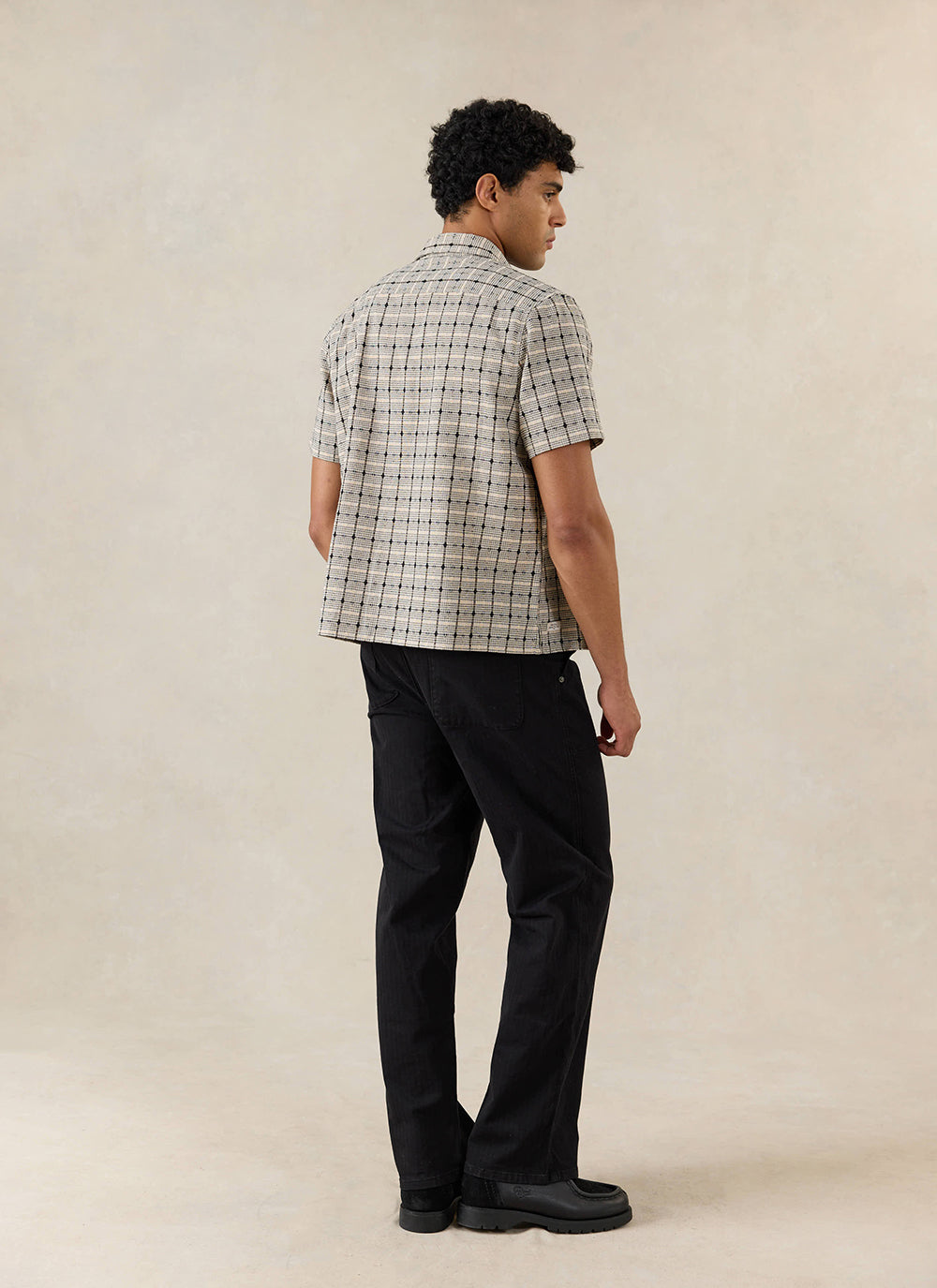 The cream textured Percival Bayley Textured Check Shirt, seen from behind and styled with black trousers in a studio setting.