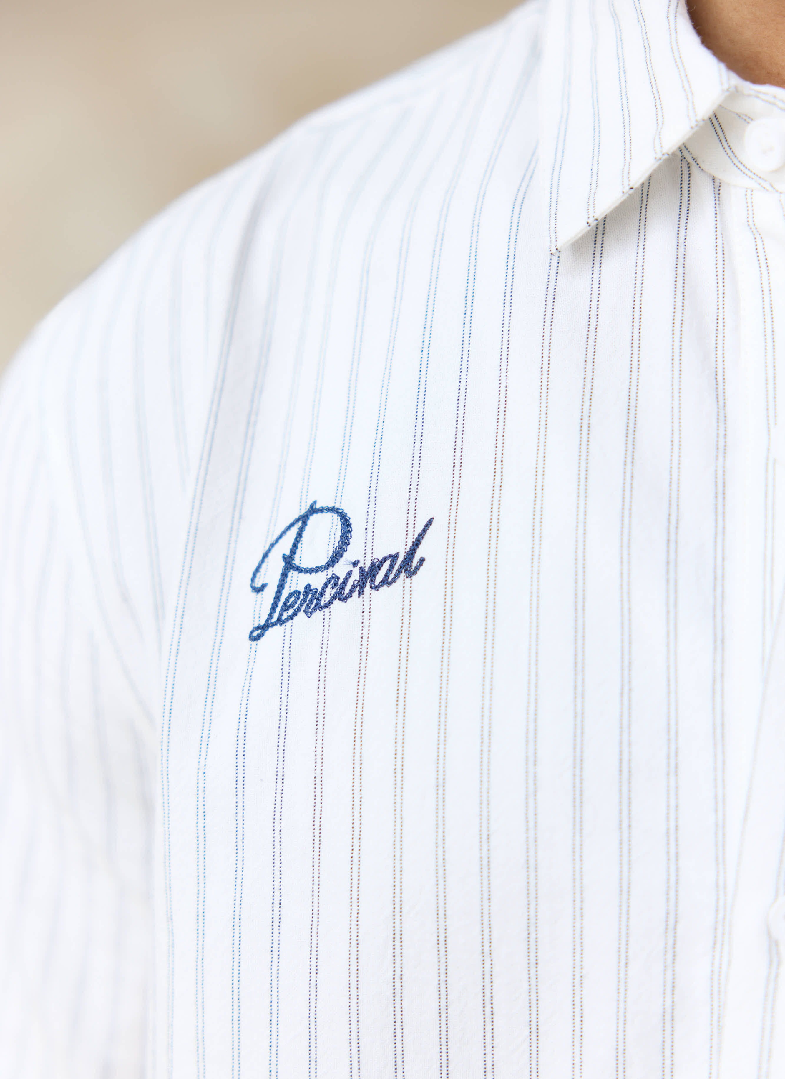 Close-up of the blue script Percival embroidery on the chest of the cream stripe cotton Lucia shirt.