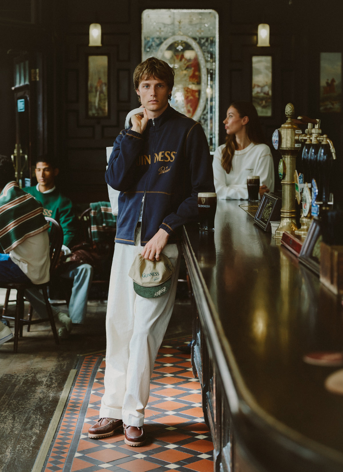 Model wearing the Guinness x Percival navy funnel neck zip up sweatshirt and white trousers inside a traditional pub.