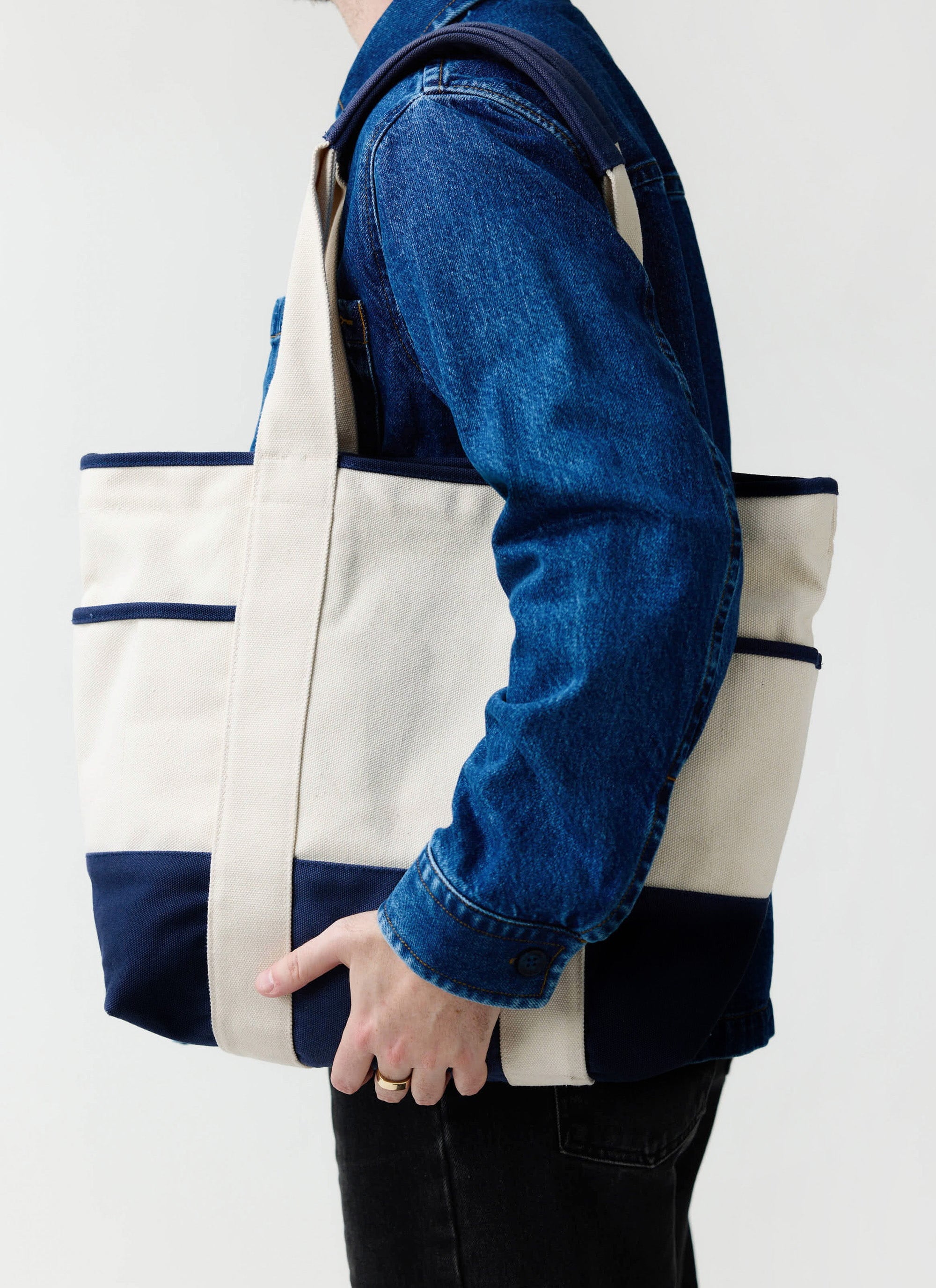 A model wearing a denim jacket carries the Percival Tote Bag in ecru and navy canvas over his shoulder.