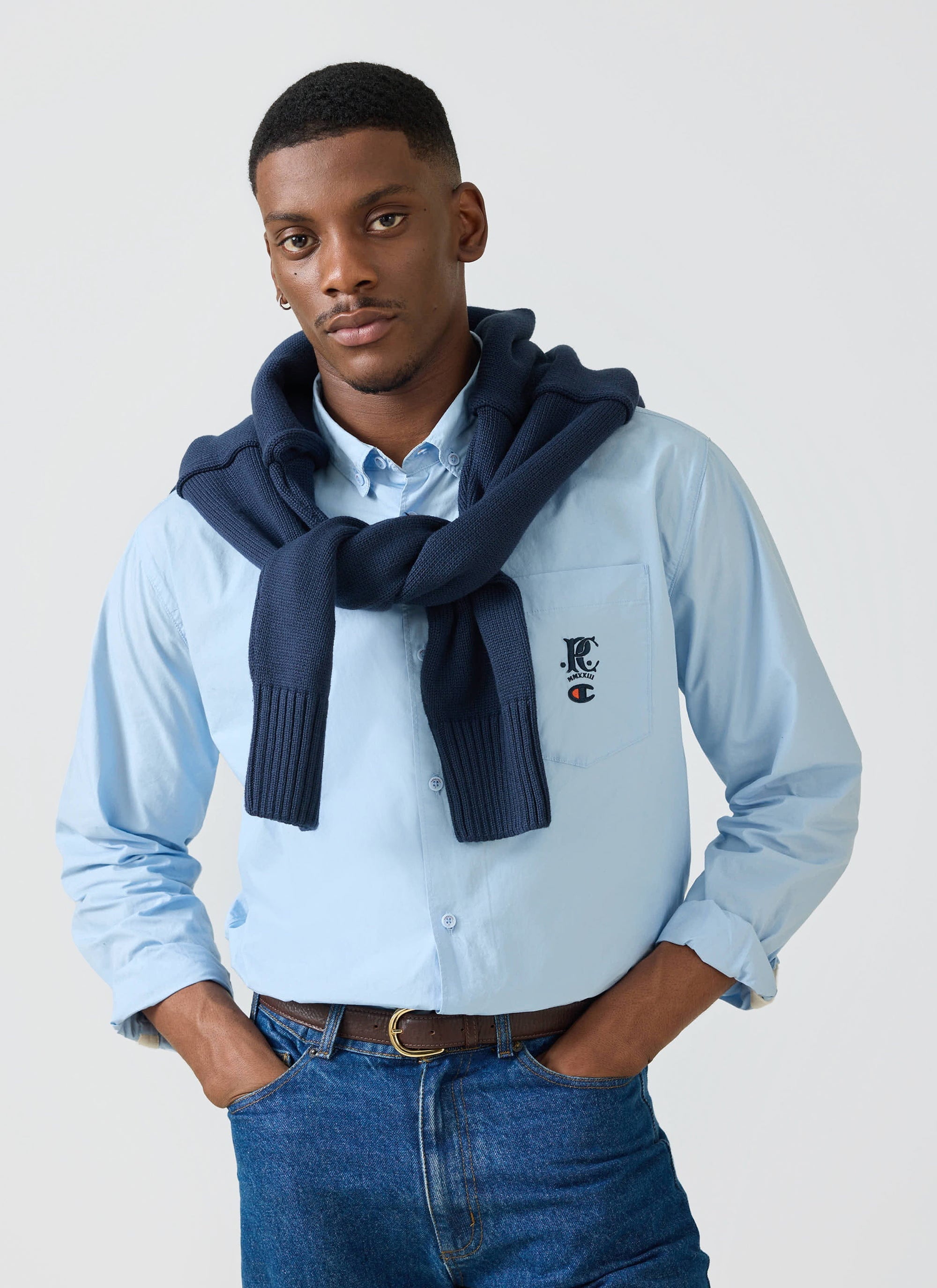 A man models the Champion x Percival Relaxed Oxford Shirt in light blue, with a navy jumper draped over his shoulders.