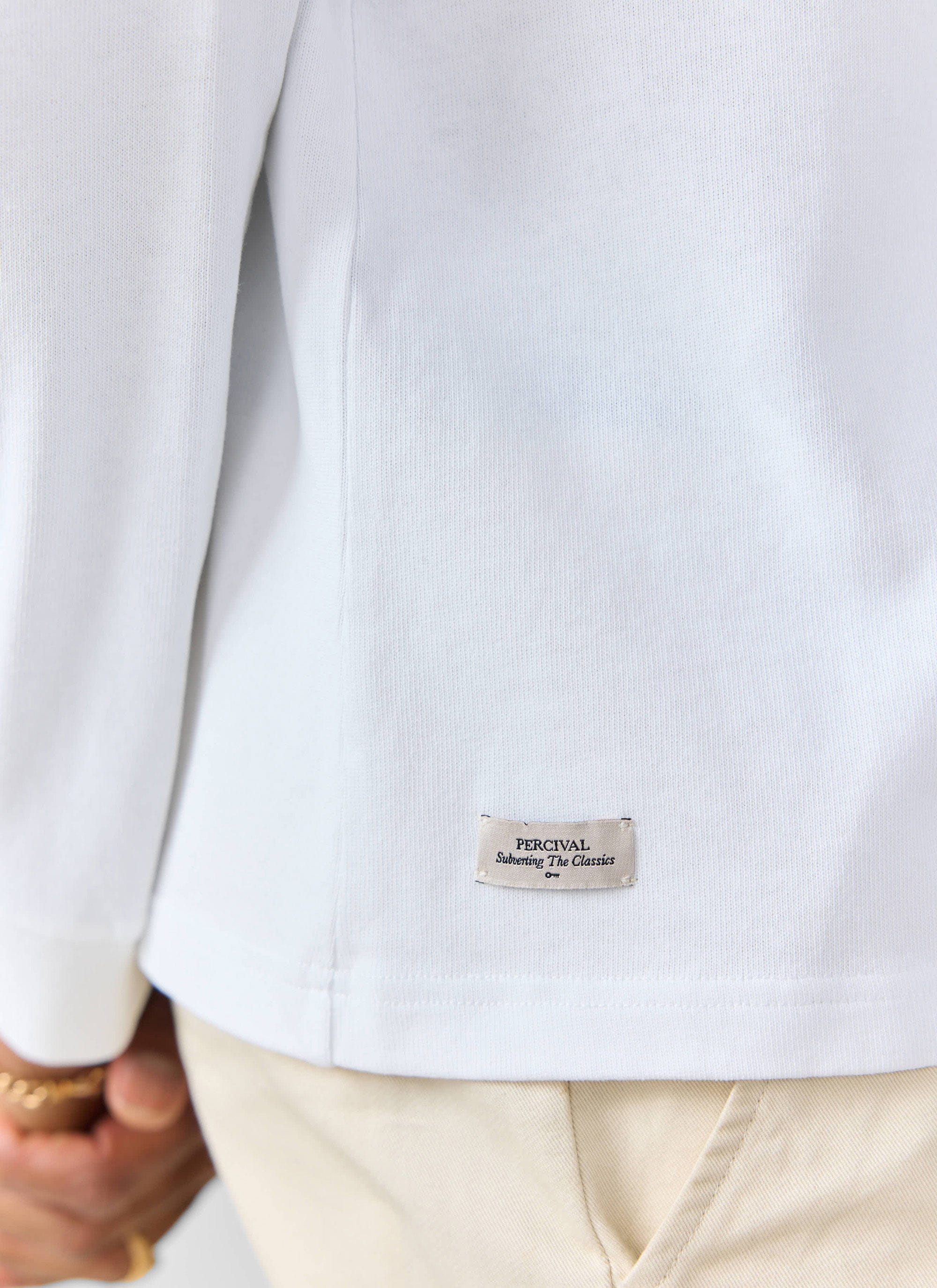 A close-up of the Percival white long-sleeve t-shirt, showing the 'Subverting The Classics' woven label at the hem.