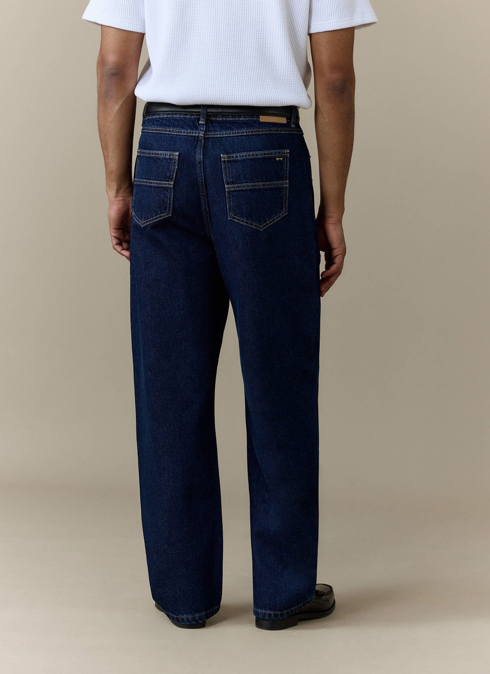 Rear view of a model wearing the Percival Pleated Denim Jeans in dark indigo blue, styled with a white t-shirt.