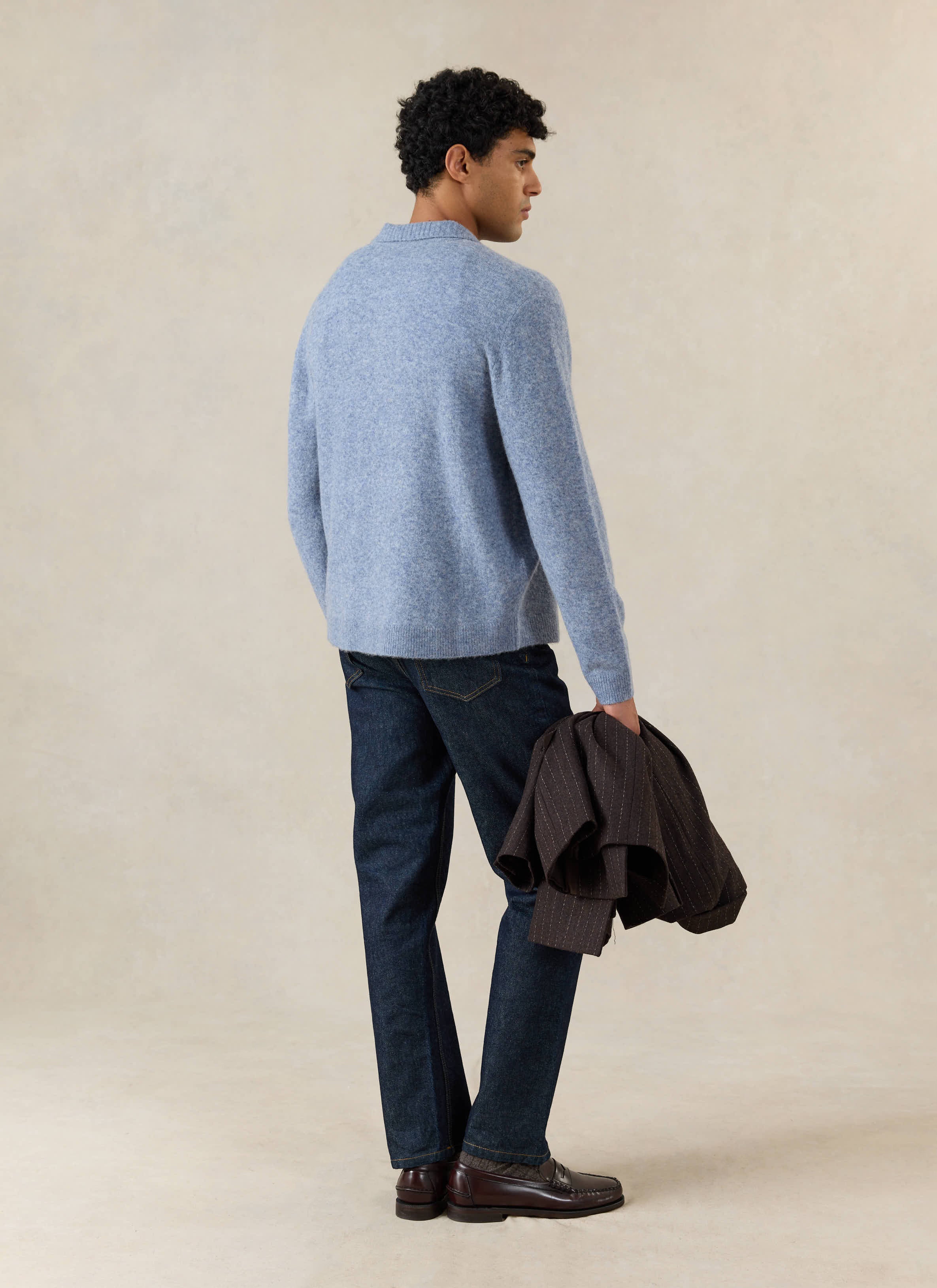 A model wears the Percival heather blue yak wool cardigan from the back, styled with dark blue jeans and holding a jacket.