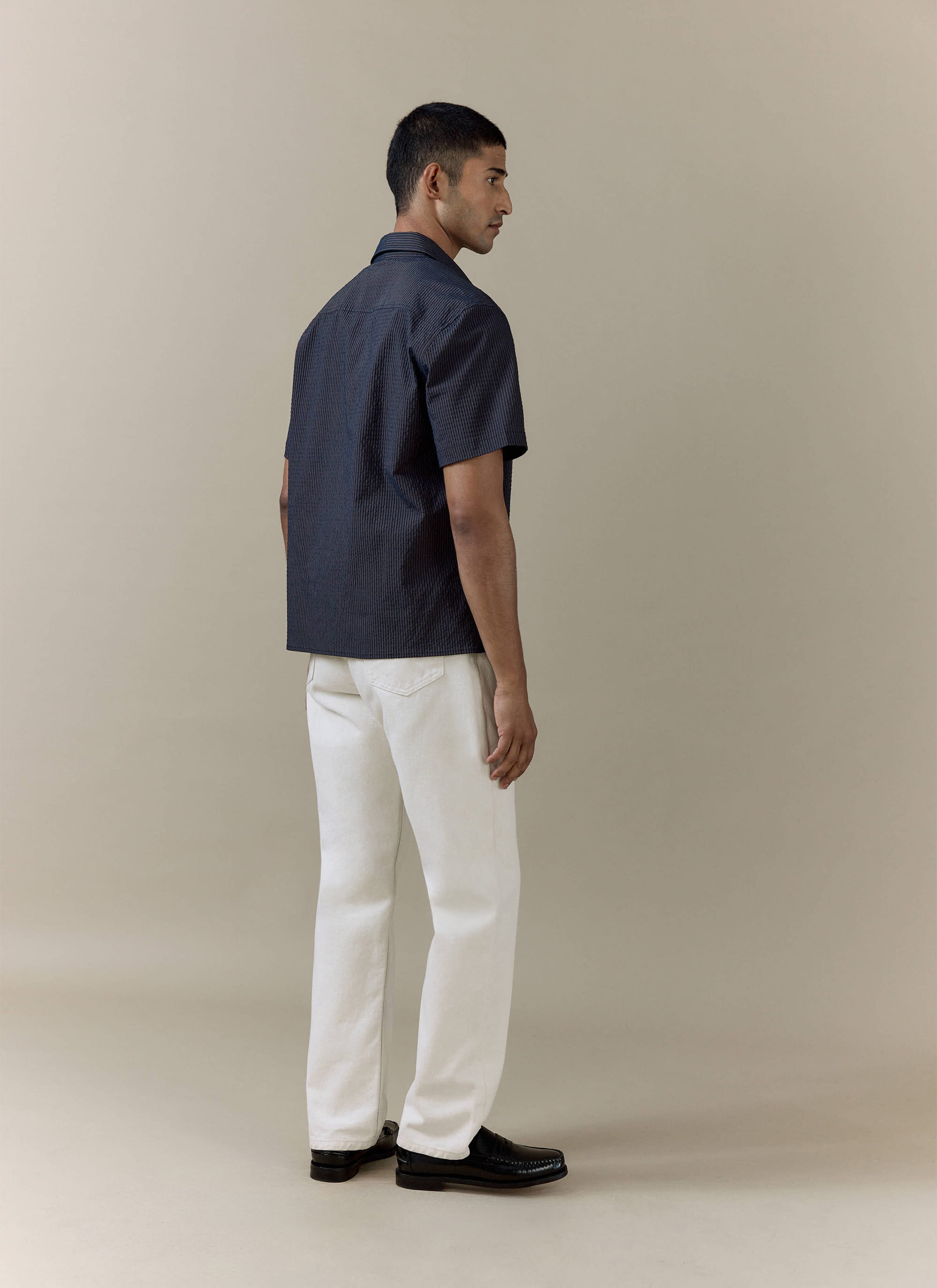 Percival Rowan Seersucker Boxy Shirt in navy stripe, shown from the back with white trousers and black loafers.