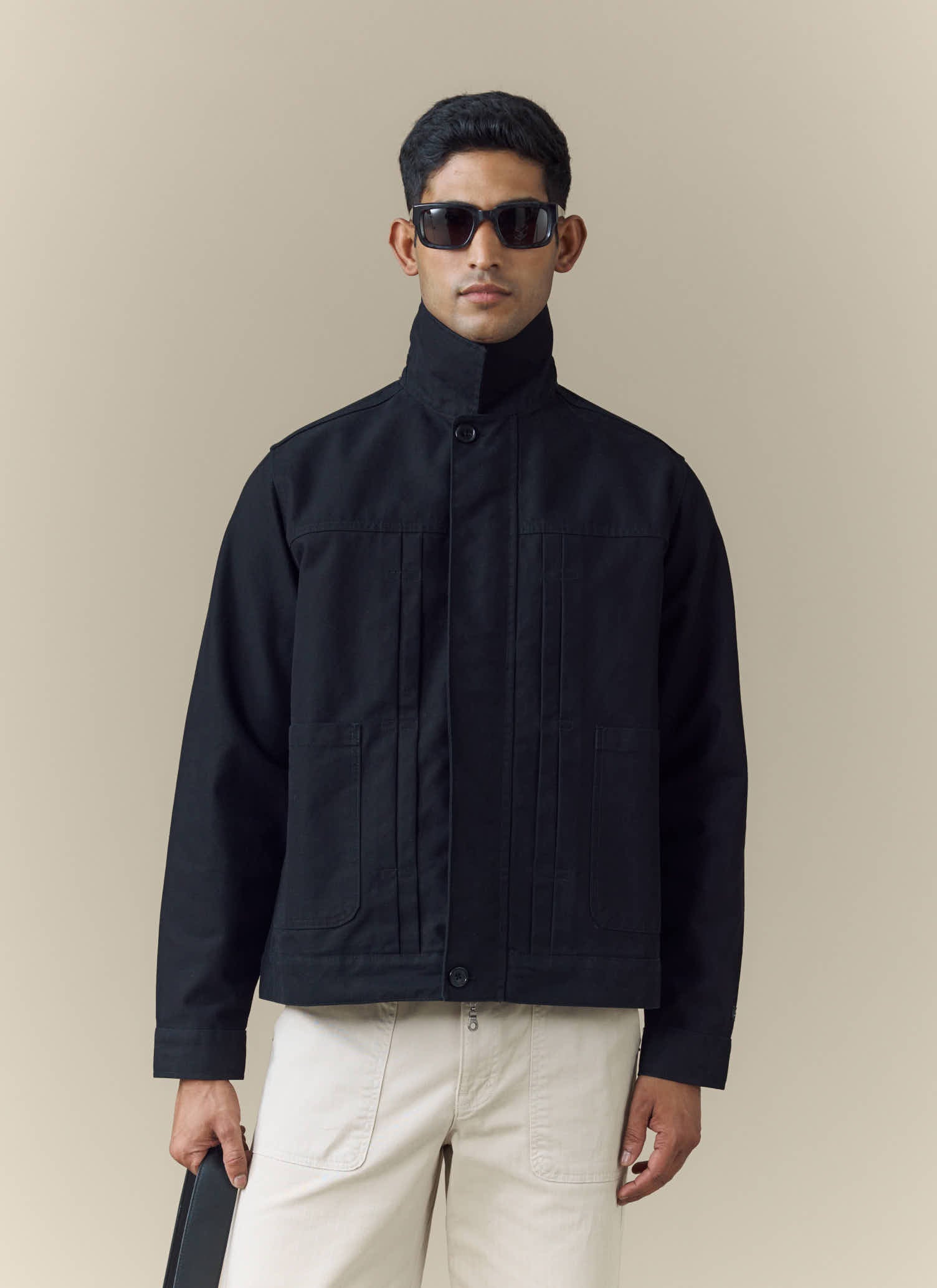A model wears the black Ernst Jacket from Percival with a high buttoned collar, sunglasses, and light-coloured trousers.