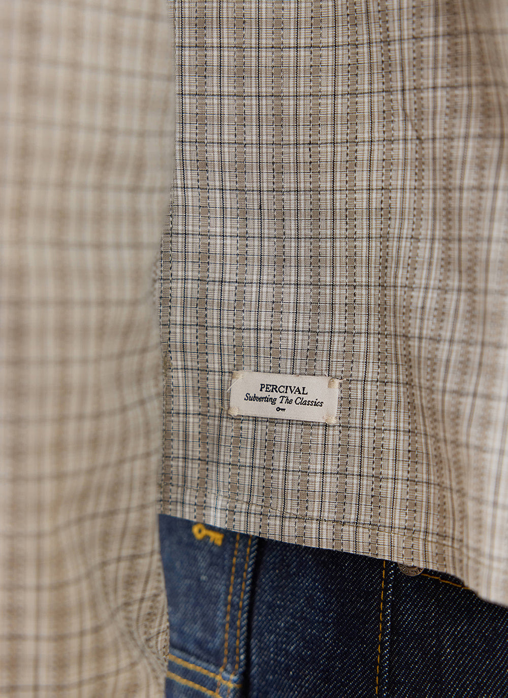 A close-up of the cream check Hinsley shirt by Percival, highlighting the branded hem tag and dark denim jeans.