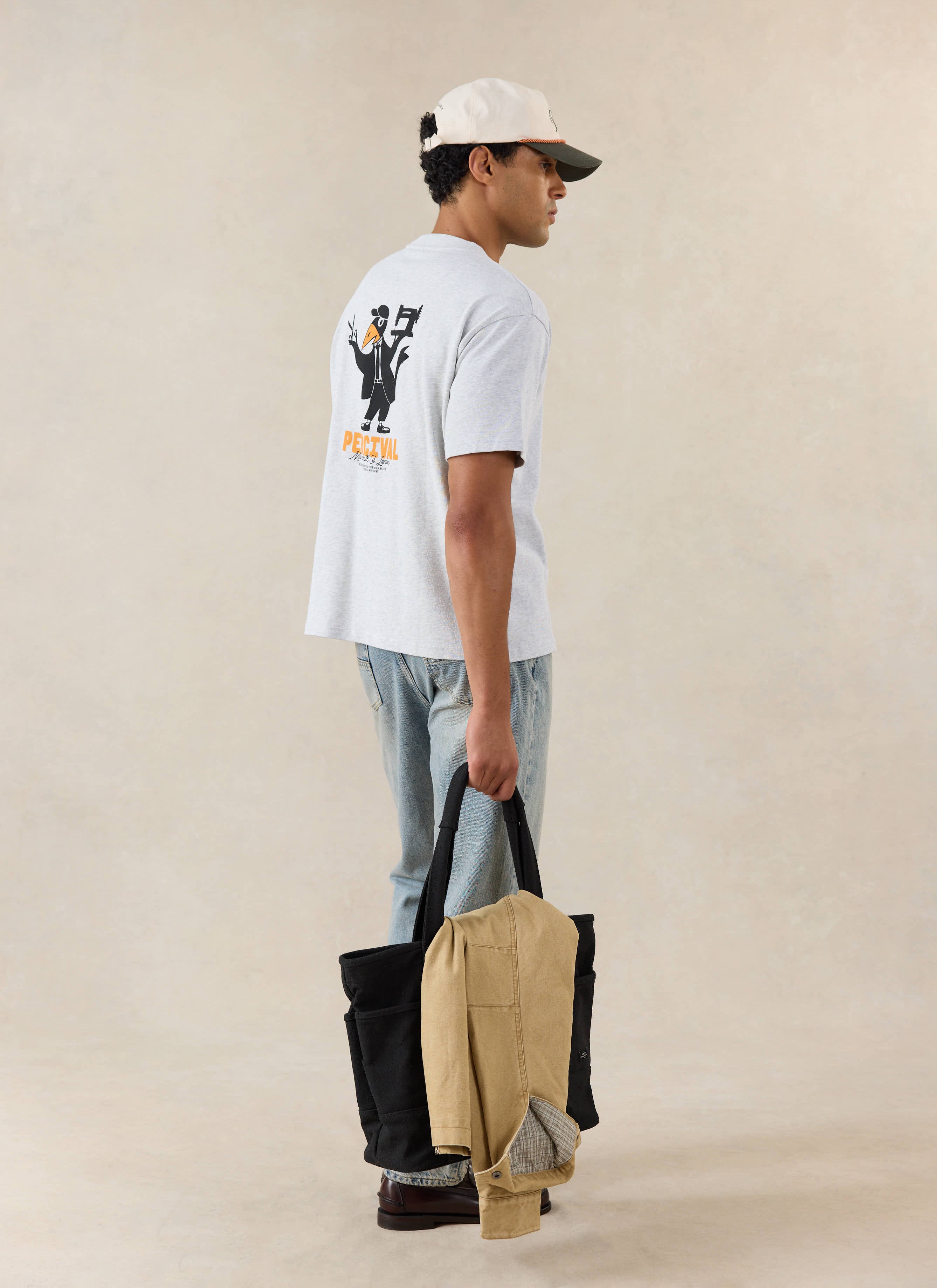 A model wears the athletic grey Percival Atticus T-shirt with a back graphic, styled with denim jeans and a tote bag.