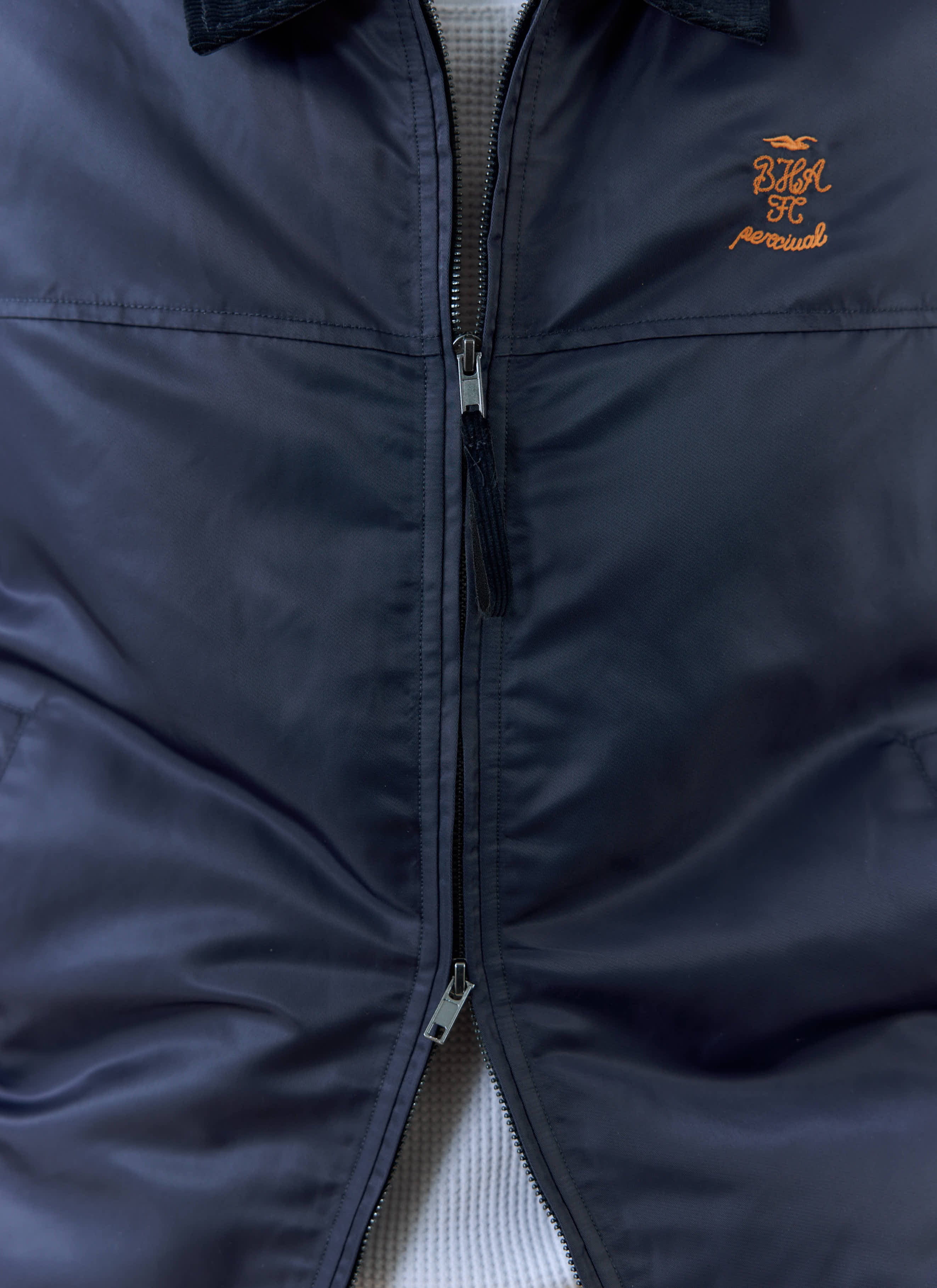 A close-up of the zip and orange chest embroidery on the navy Percival x Brighton & Hove Albion F.C. Coach Jacket.