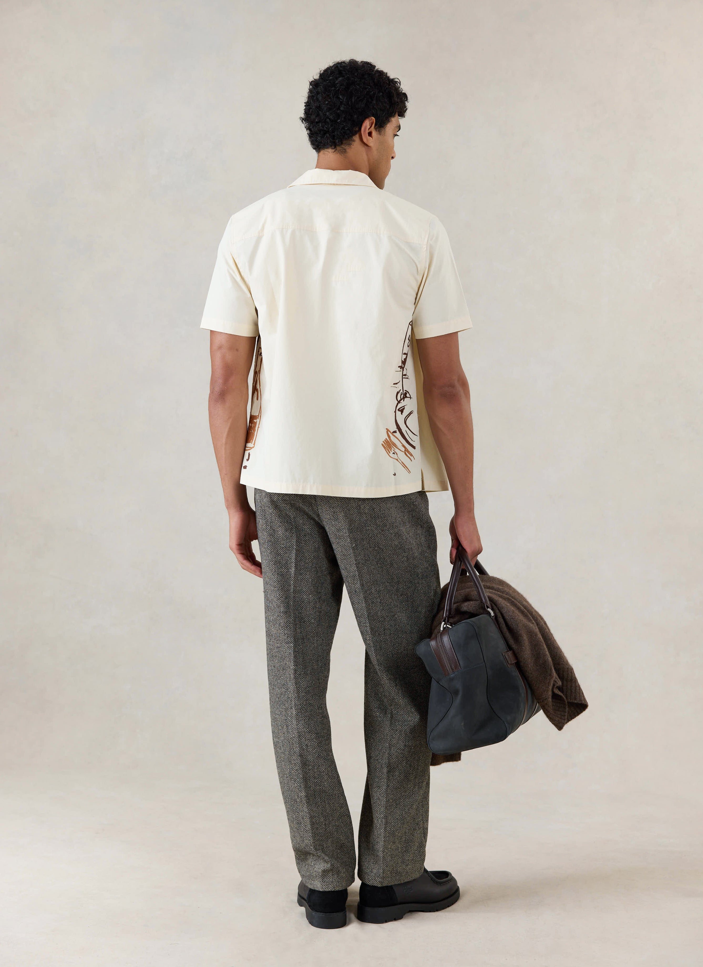 Rear view of a model in the cream Percival Cuban shirt, styled with textured trousers and holding a bag.
