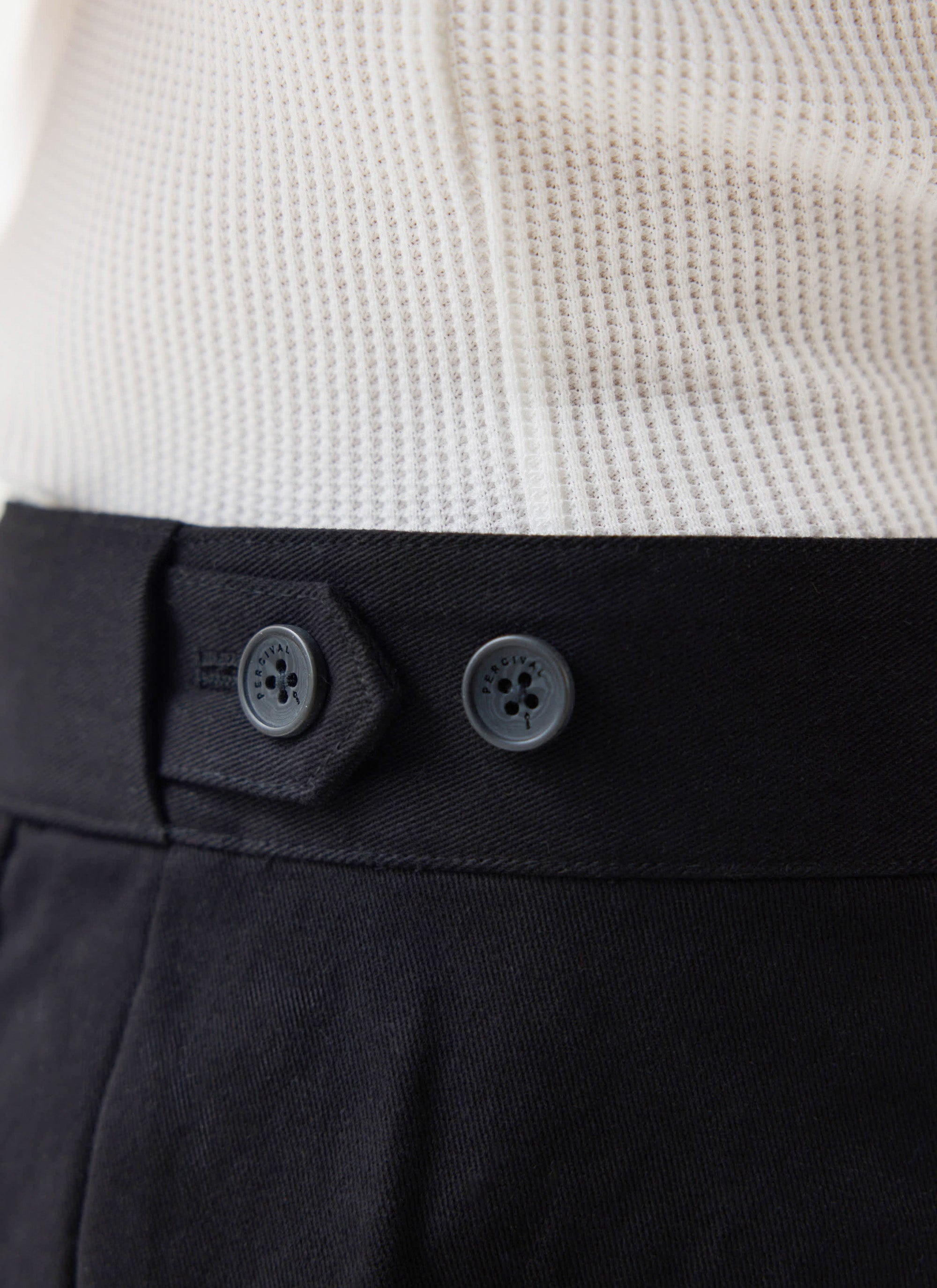 A close-up of the branded buttons and waistband of the black cotton Straight Leg Chino from Percival.
