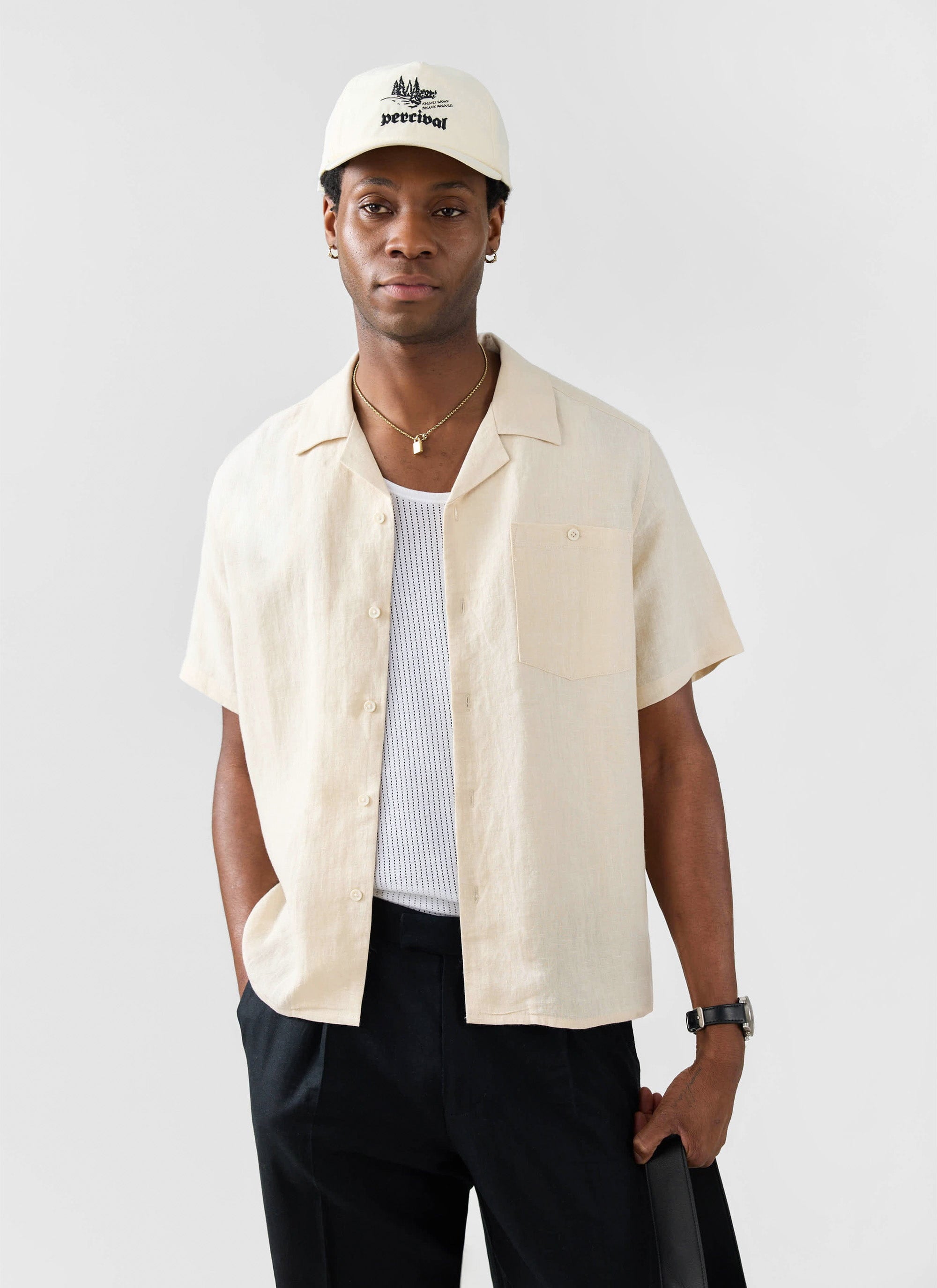 A model wears the Percival cream linen Short Sleeve Cuban Shirt with black trousers and a Percival baseball cap.