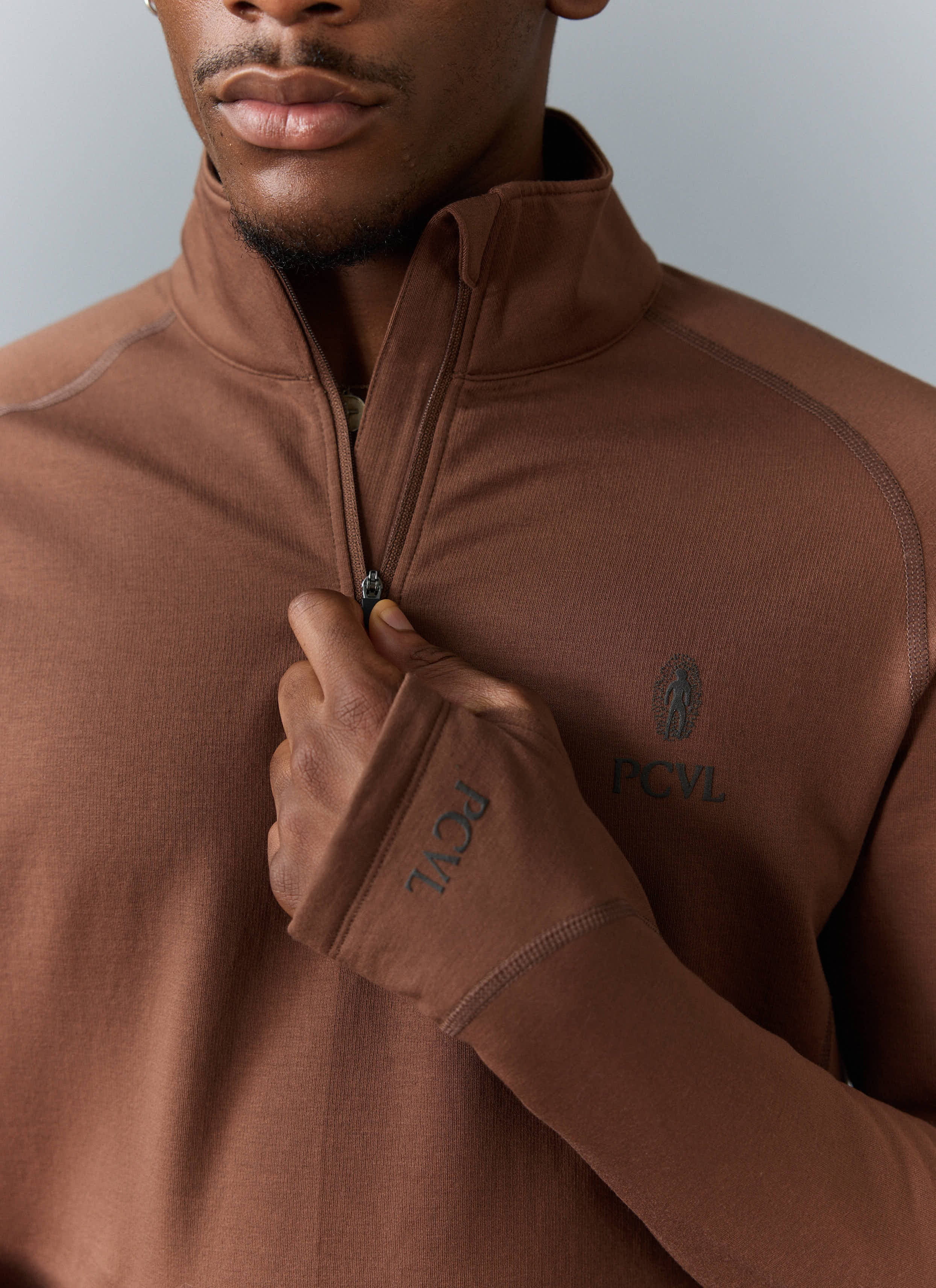 A close-up of a model wearing the Percival PCVL Base Layer Quarter Zip in rust, showing the zip, chest logo and cuff detail.