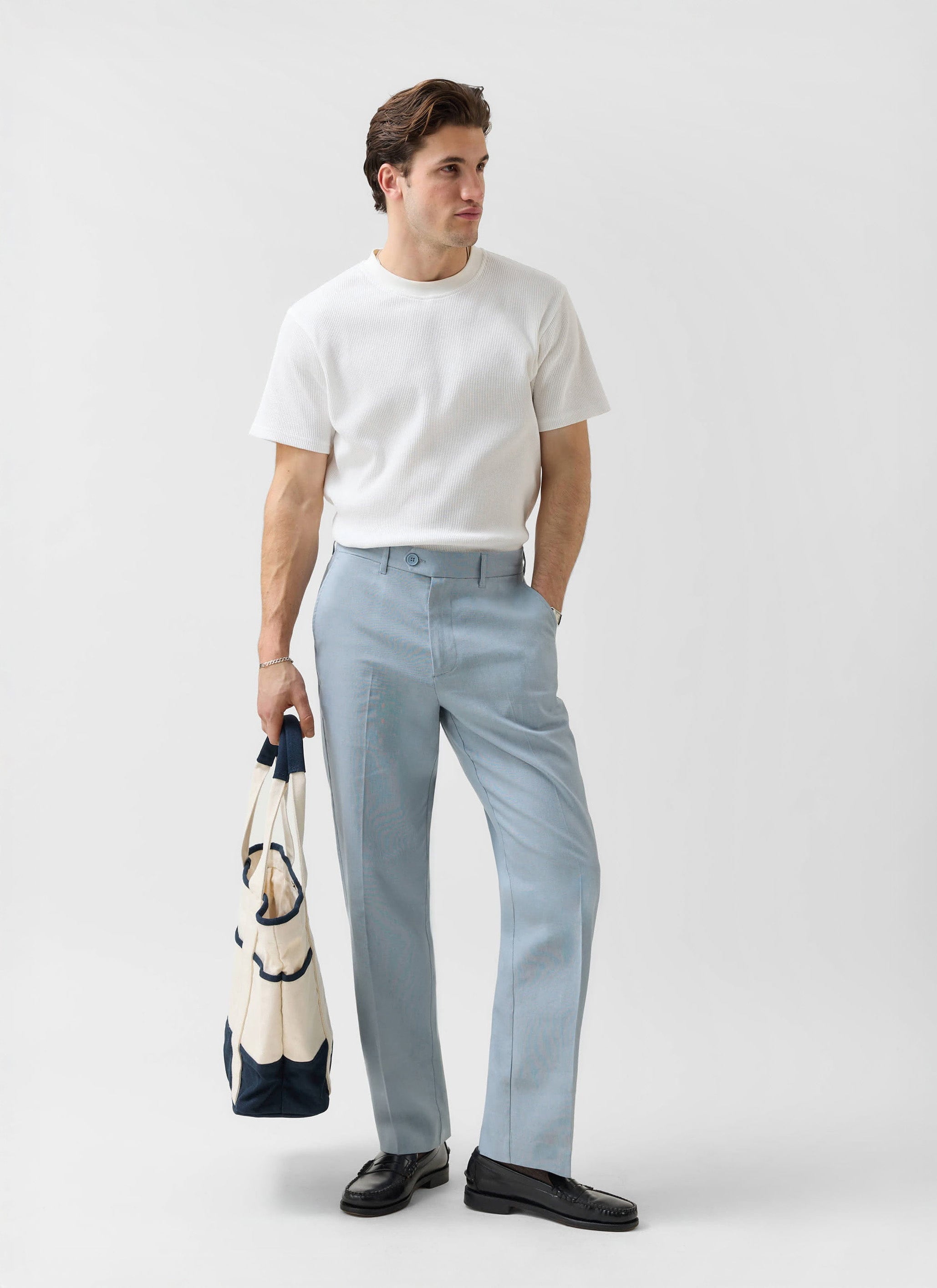 Model wears Percival Menswear light blue tailored linen trousers with a white t-shirt and black loafers, holding a tote bag.