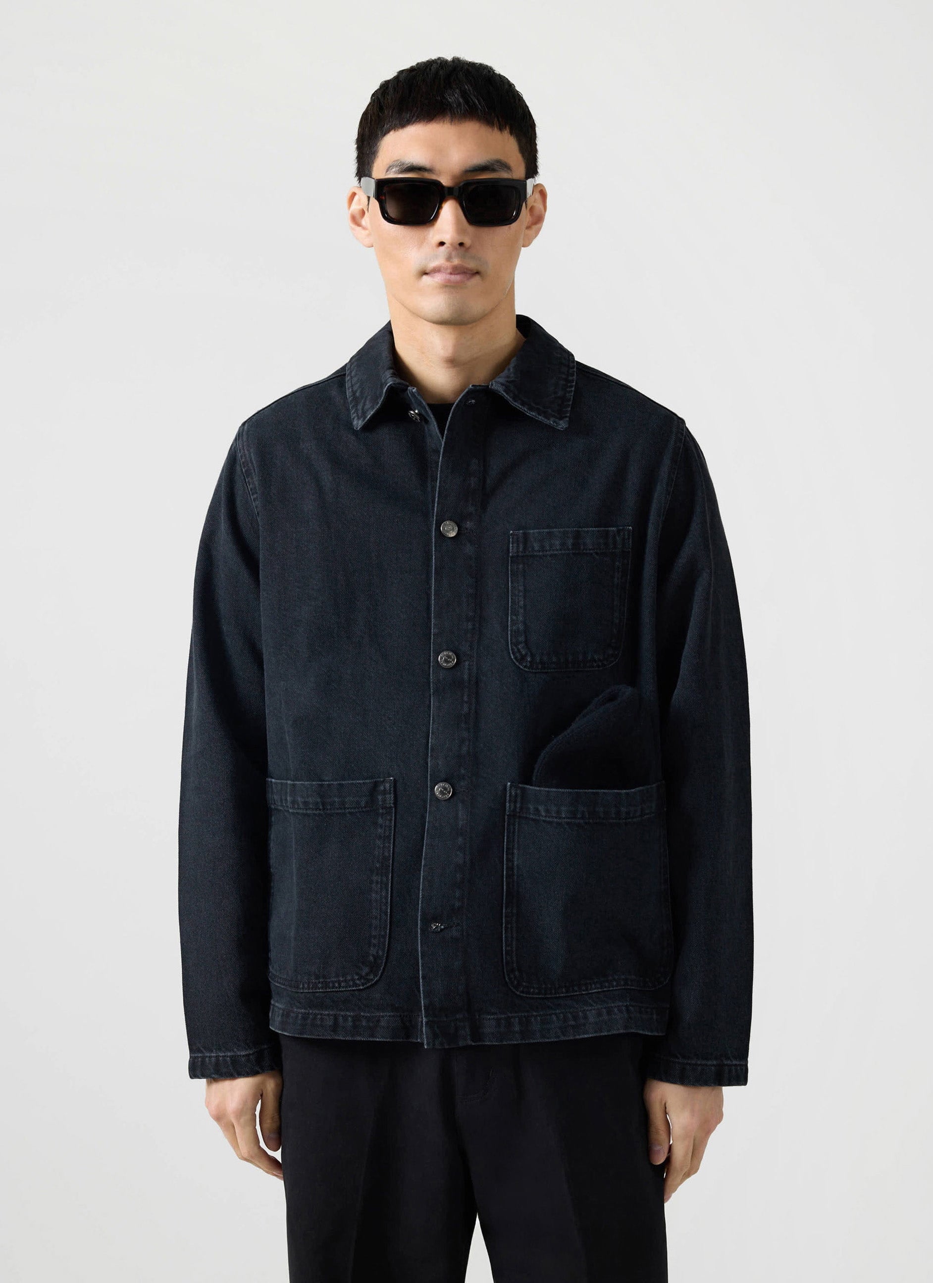 Front view of a model wearing the Percival Gove Chore Jacket in black denim, styled with black sunglasses.