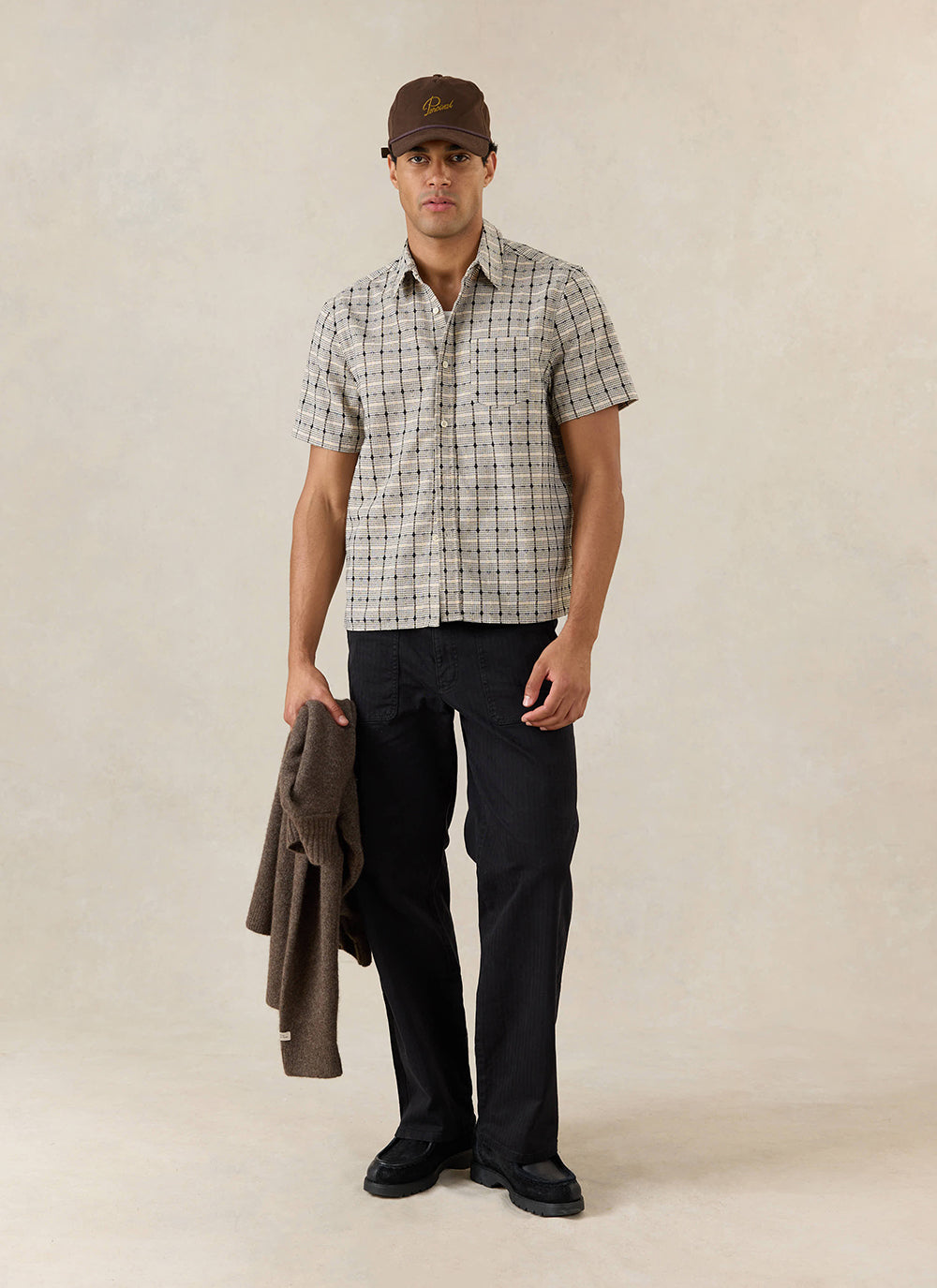 A model wears the cream Percival Bayley Textured Check Shirt, styled with black Lungo Carpenter Trousers and a brown cap.