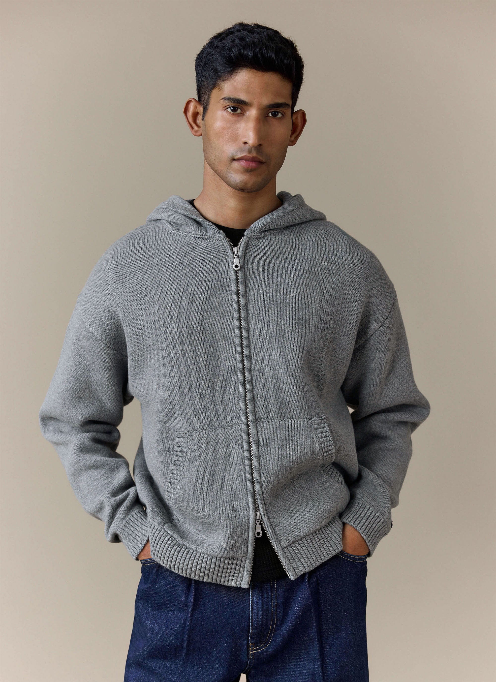 A model wears the grey Chronicle Knitted Hoodie from Percival with dark jeans, showing the zip front and kangaroo pocket.