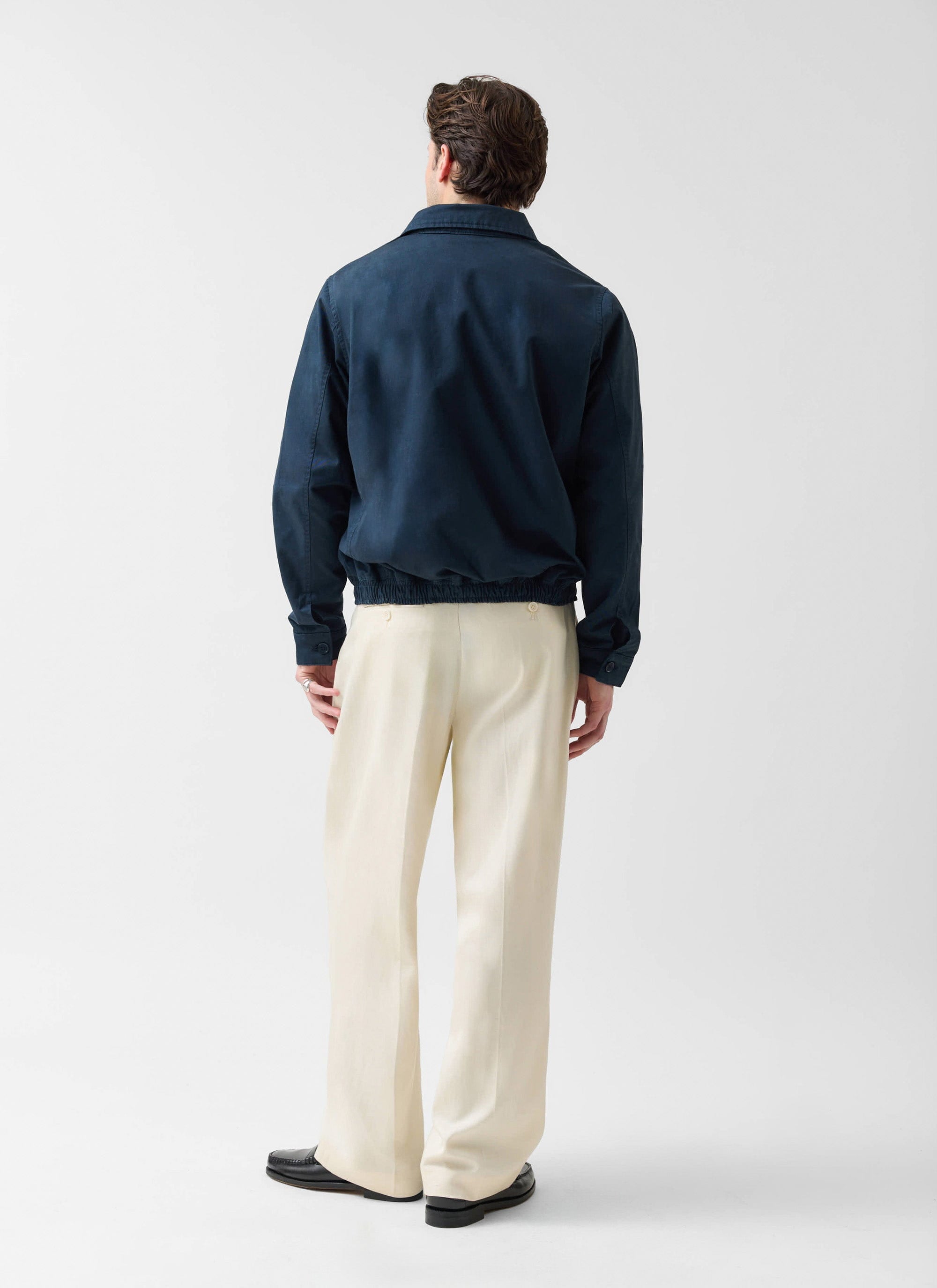 A model, seen from behind, wears the navy cotton Leo Harrington Jacket from Percival with wide-leg cream trousers.