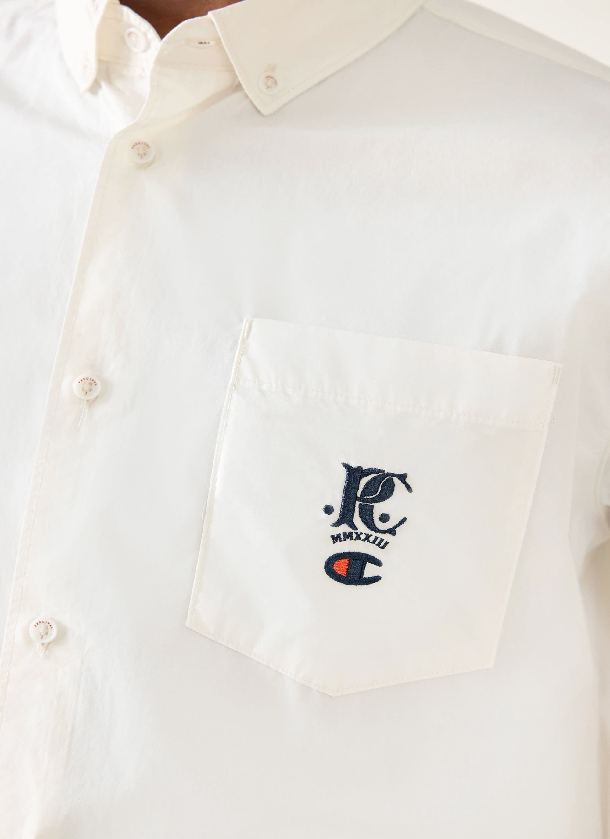 A close-up of the Champion x Percival embroidery on the chest pocket of the ecru Oxford shirt from Percival.