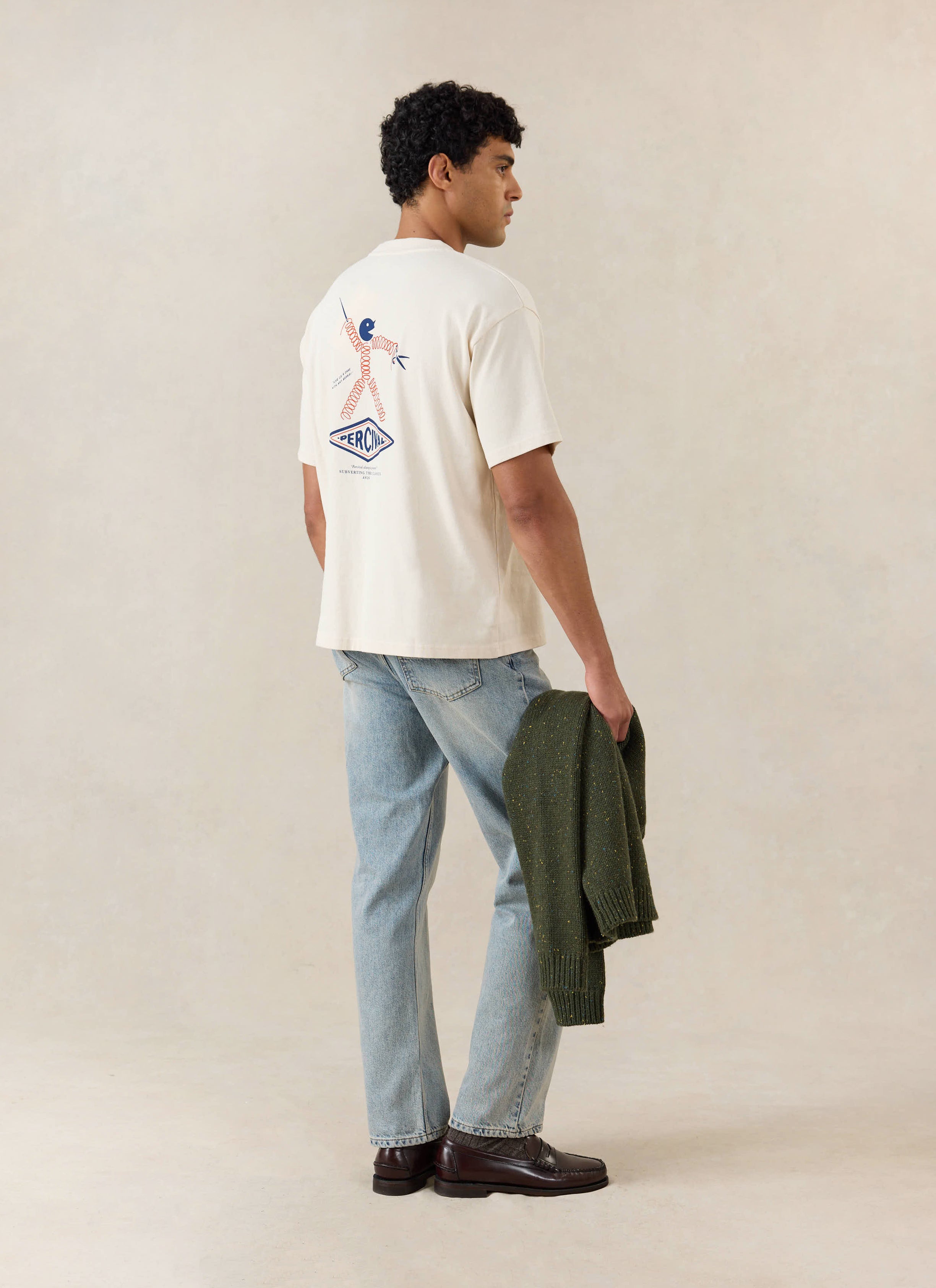 A model from behind wears the cream Percival Kingston boxy t-shirt with a back graphic, styled with light blue jeans.