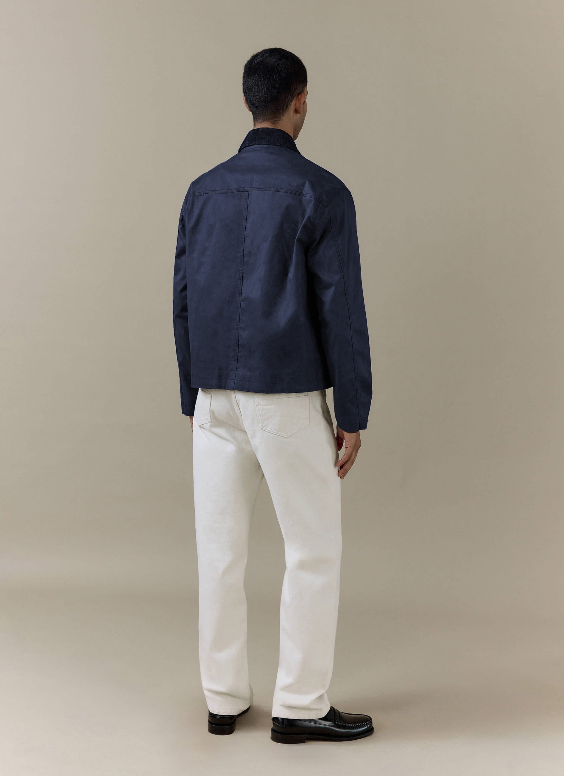 Rear view of the navy blue Percival Wax Zip Jacket in waxed cotton, styled with off-white trousers.