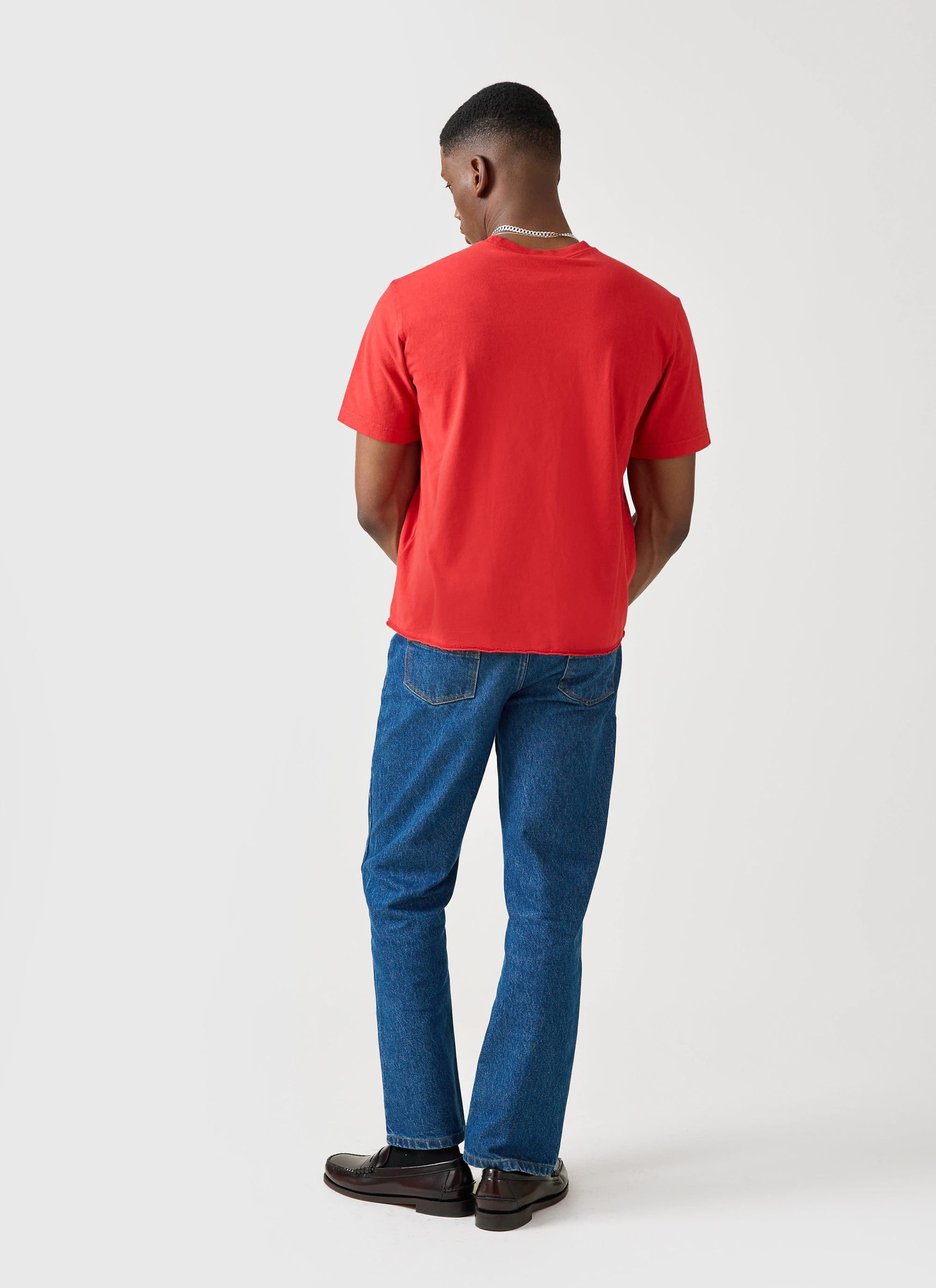 Back view of a model wearing the red Champion x Percival raw hem t-shirt, styled with blue jeans and dark loafers.