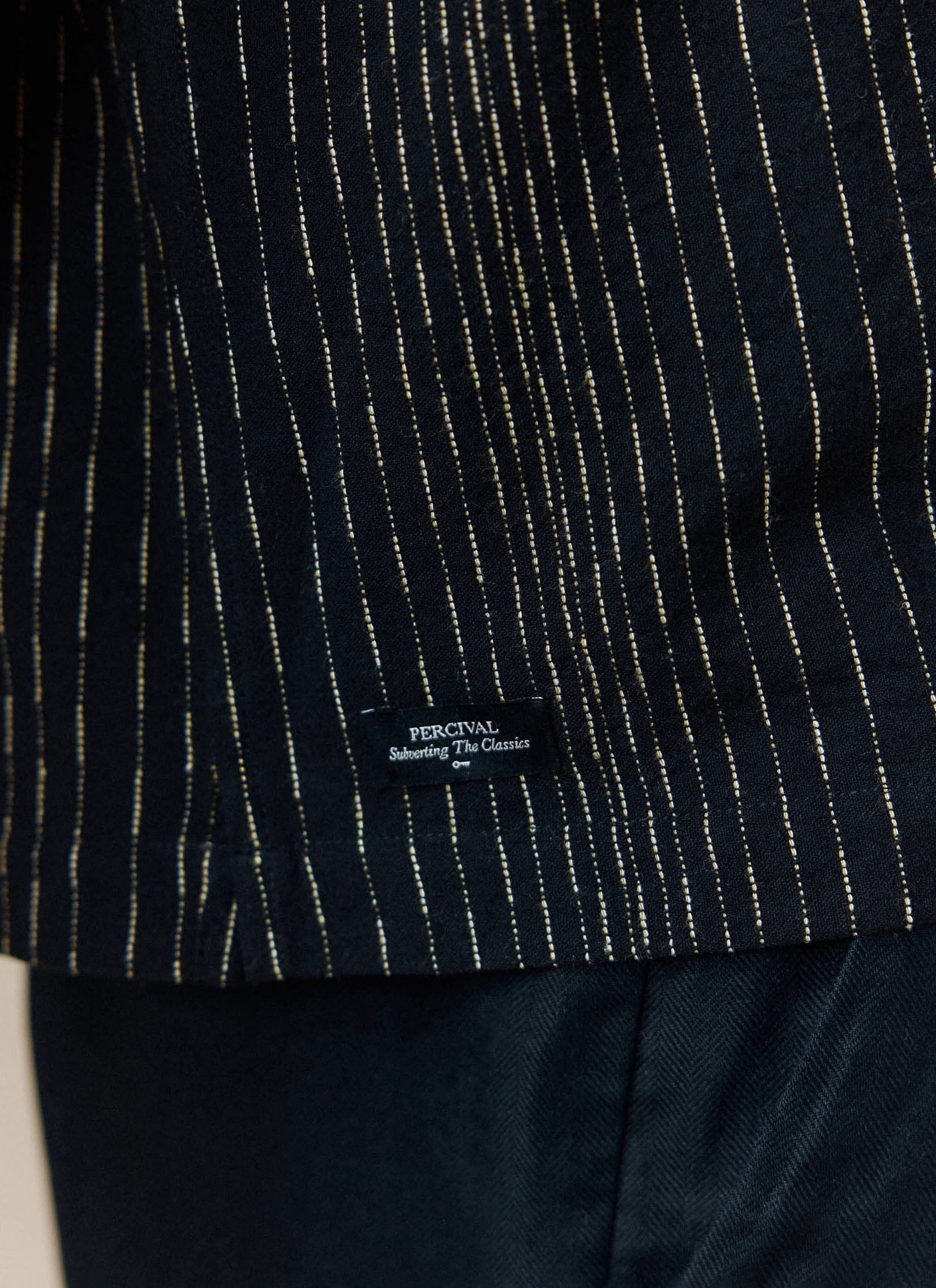 A close-up of the branded hem label on the black Leland Cuban Boxy Shirt from Percival, highlighting the pinstripe fabric.