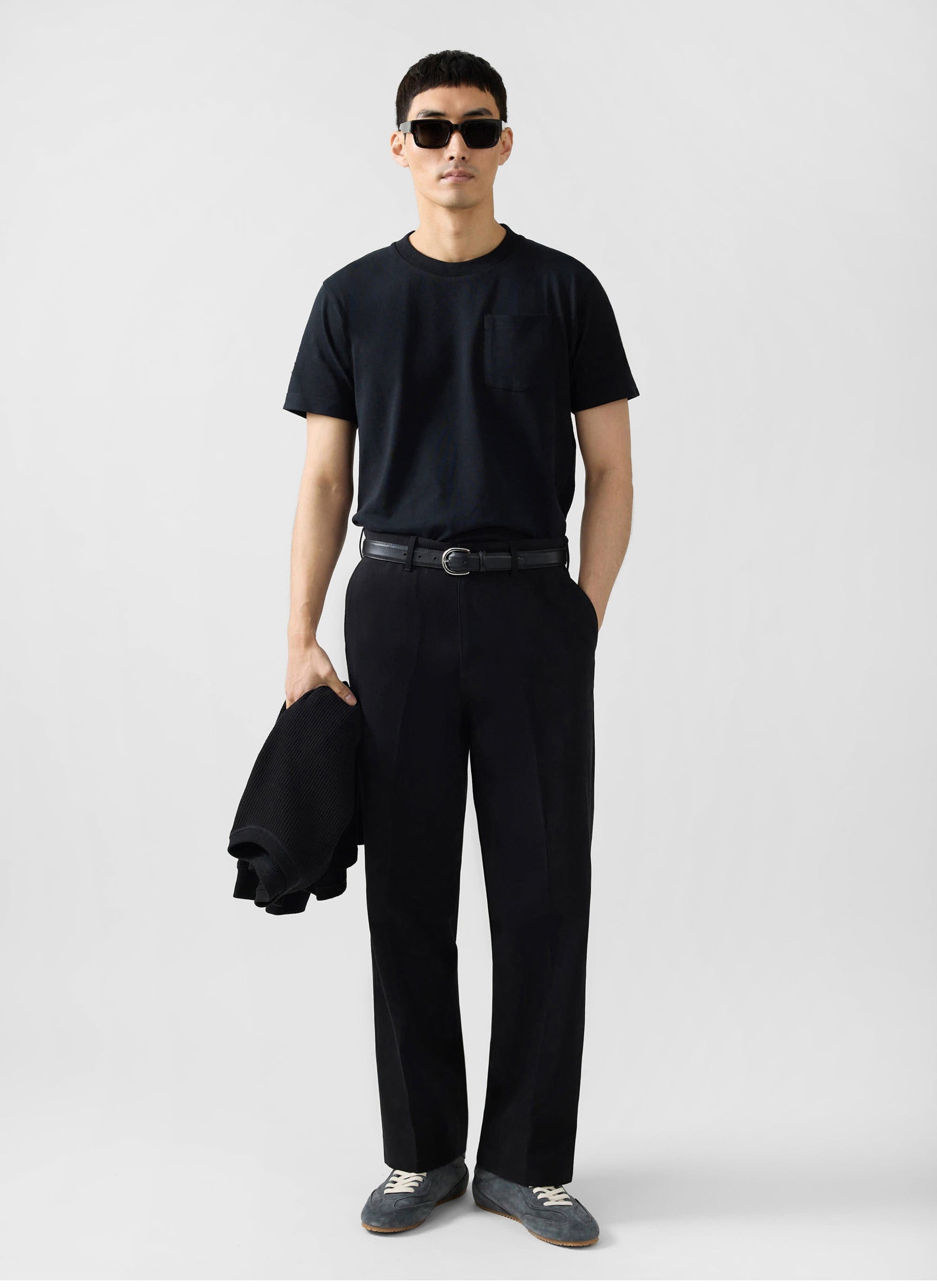 A man models the Percival black organic cotton pocket t-shirt, styled with black trousers, a belt, and sunglasses.