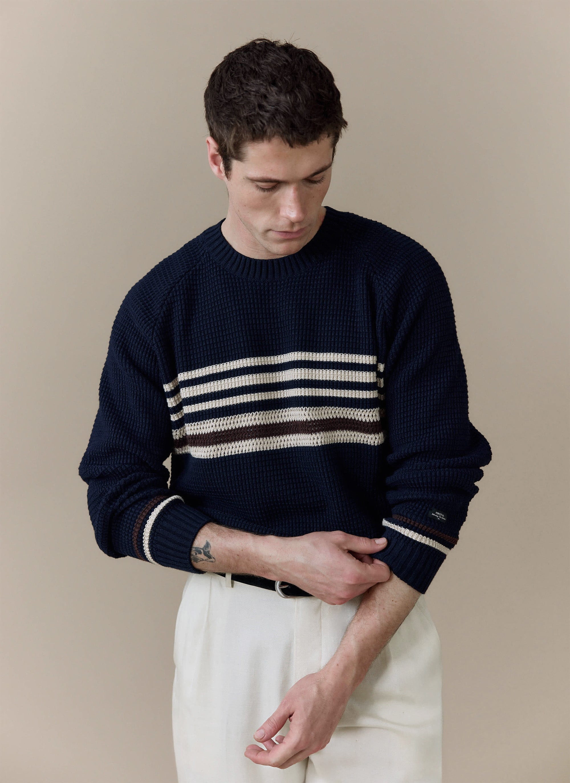 A model wears the navy Sergio Crochet Jumper from Percival, styled with white trousers against a neutral studio background.