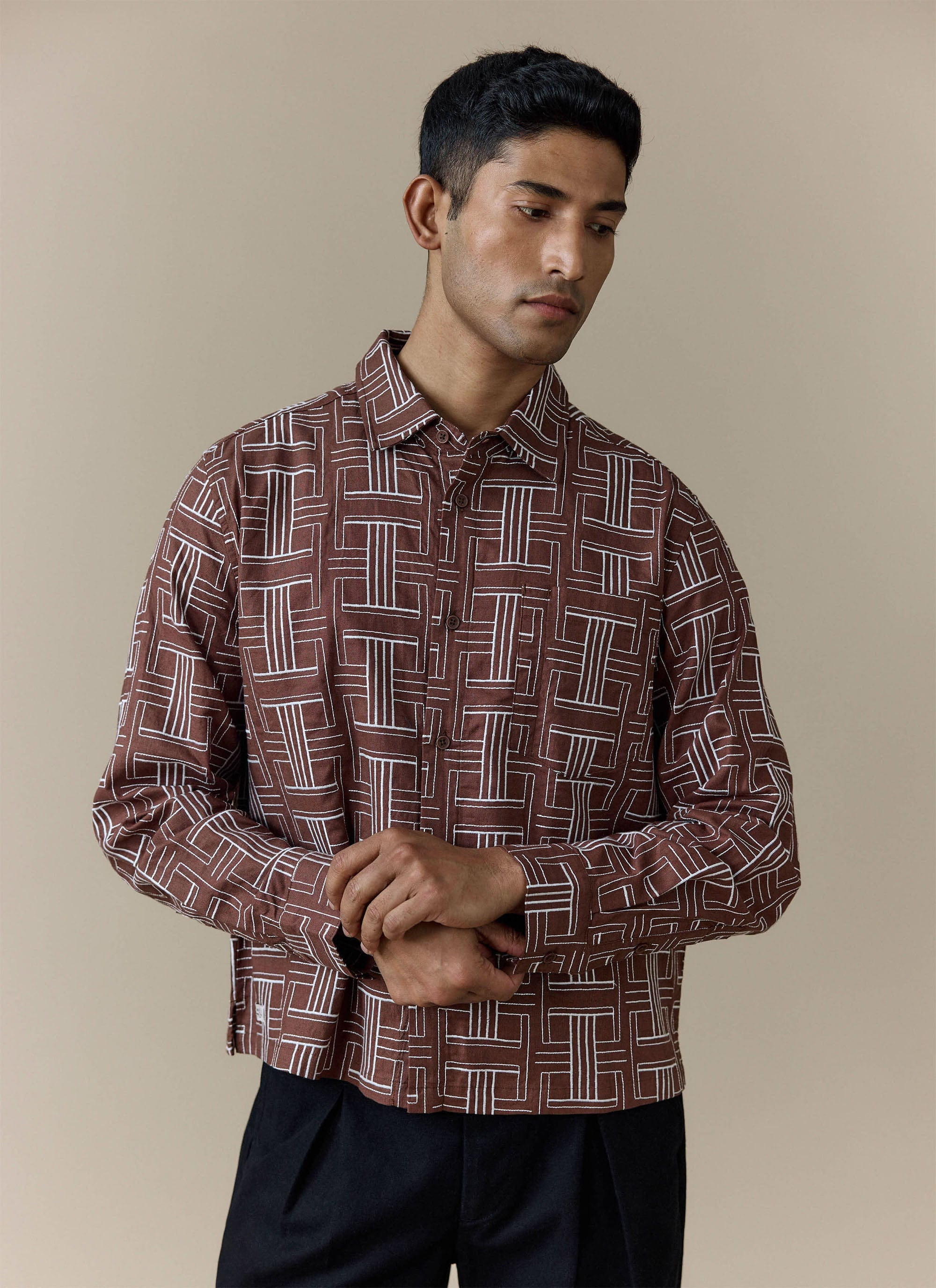 A model wears the brown Sashiko Boxy Shirt from Percival, featuring a white geometric pattern, styled with black trousers.