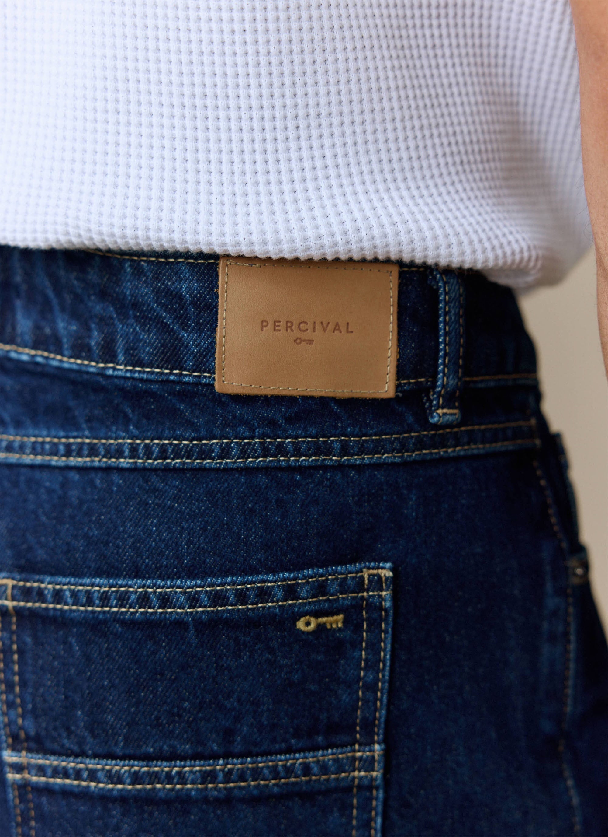 Close-up of the Percival branded tan leather patch on the waistband of the Dark Indigo Pleated Denim Jeans.