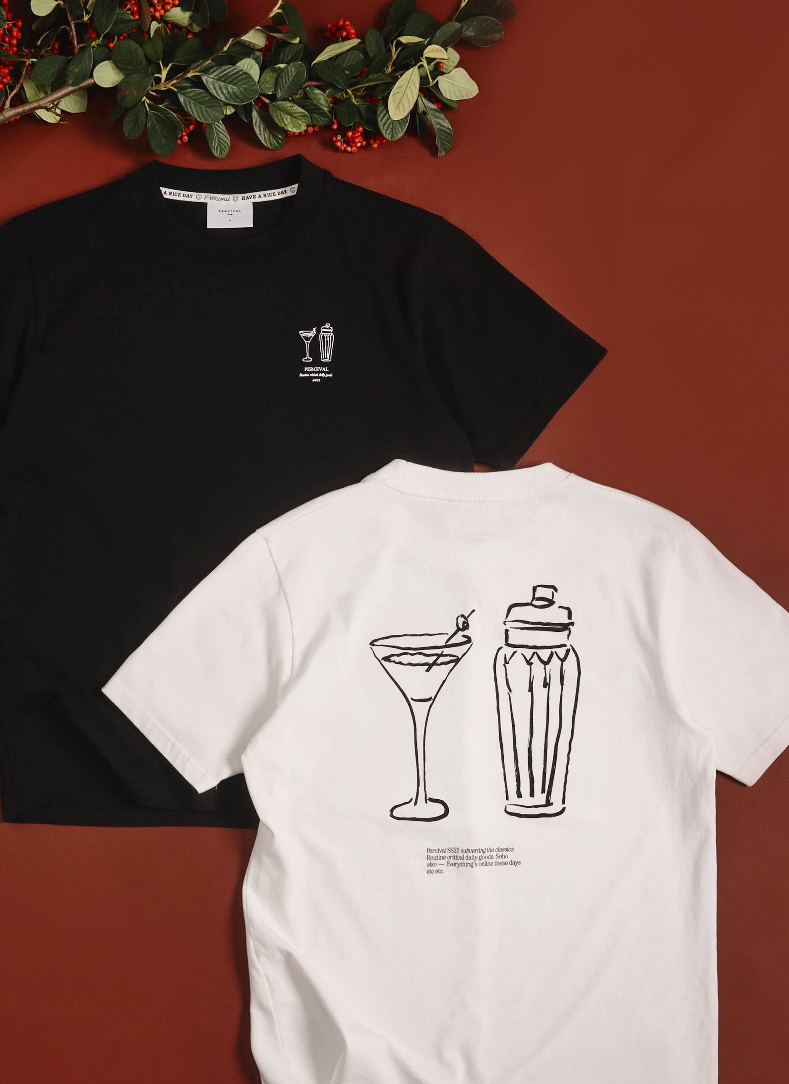 The Percival Mixologist T-shirt in black, styled with the white version, on a rich red background with a festive branch.