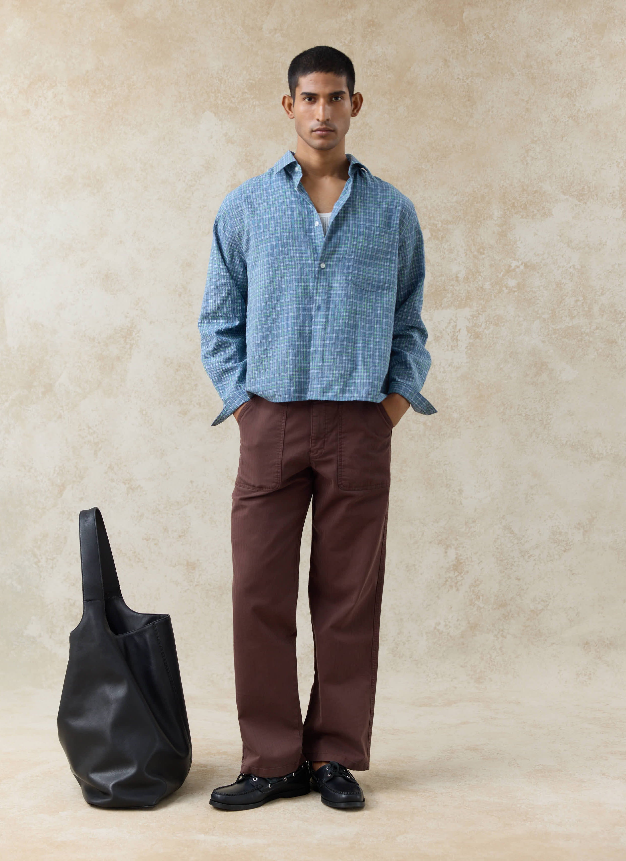 A model wears the blue Percival Hinsley Checked Boxy Shirt in textured cotton, paired with brown utility trousers.