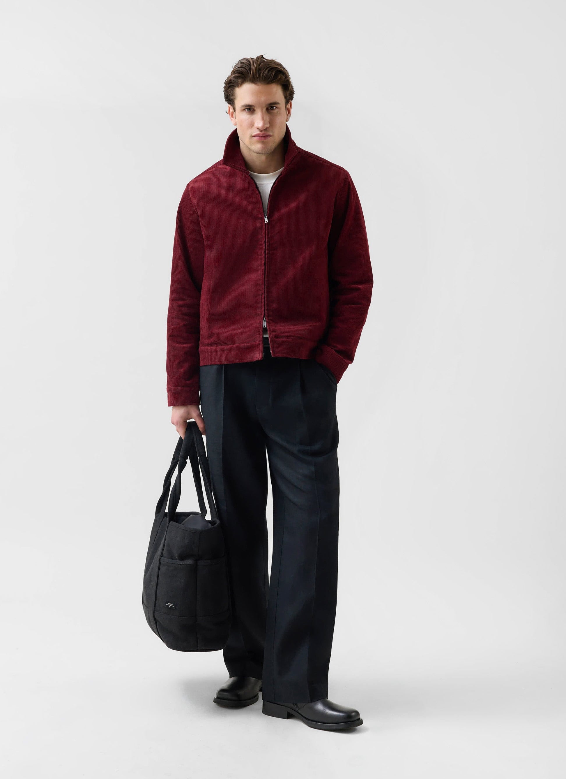 A model wears the Percival Marcel Cord Jacket in burgundy, styled with dark trousers and holding a black tote bag.