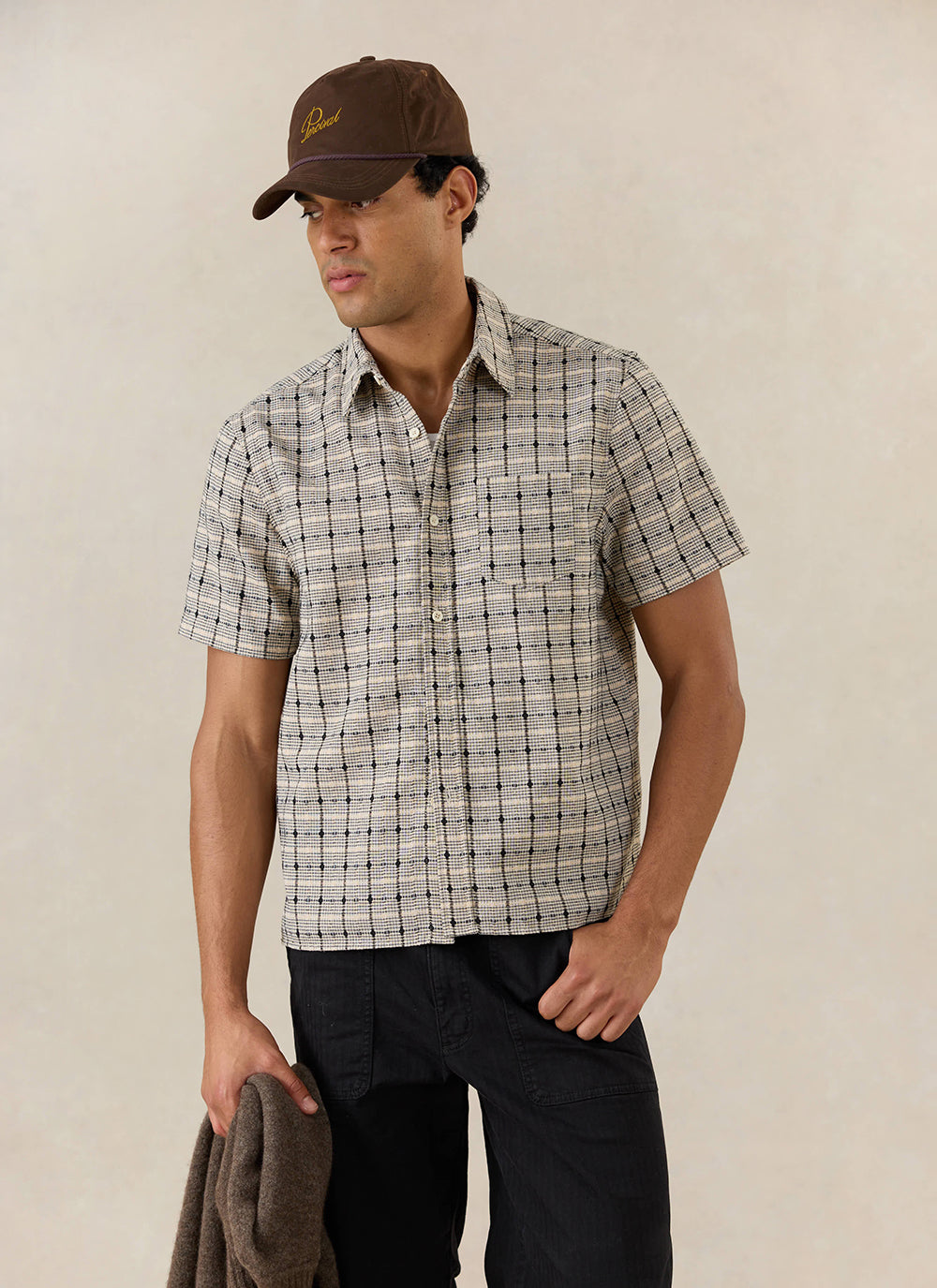 A model wears the cream Percival Bayley Textured Check Shirt, styled with a brown cap and black trousers.