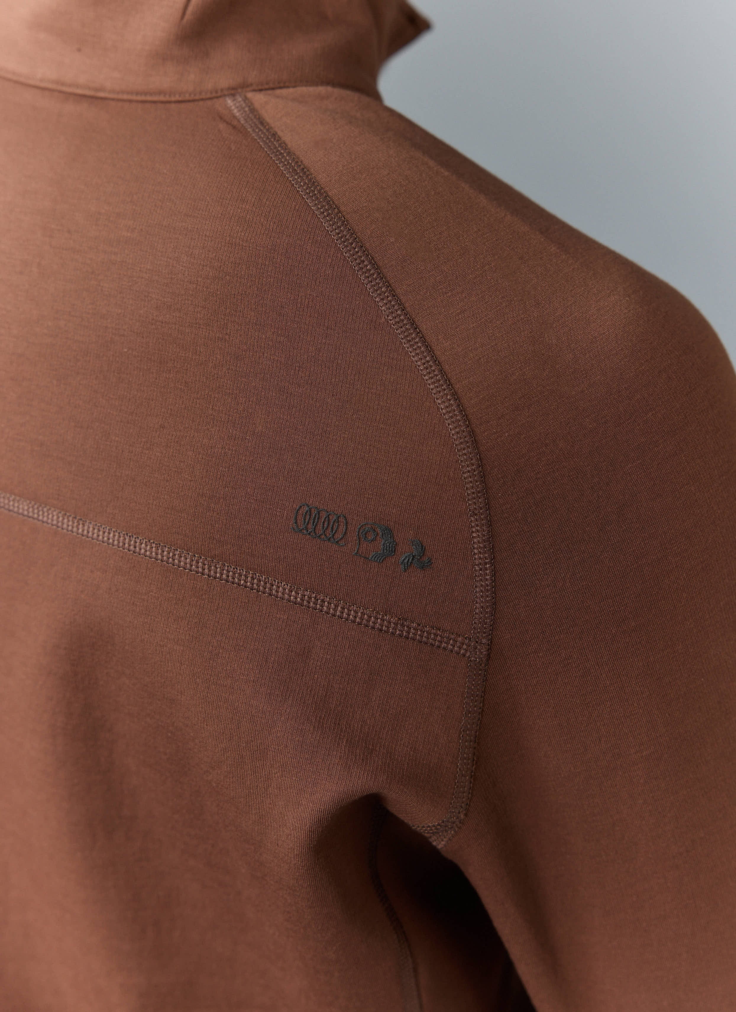 A close-up of the Percival PCVL Base Layer Quarter Zip Jacket in rust, showing the chest logo and raglan sleeve detail.