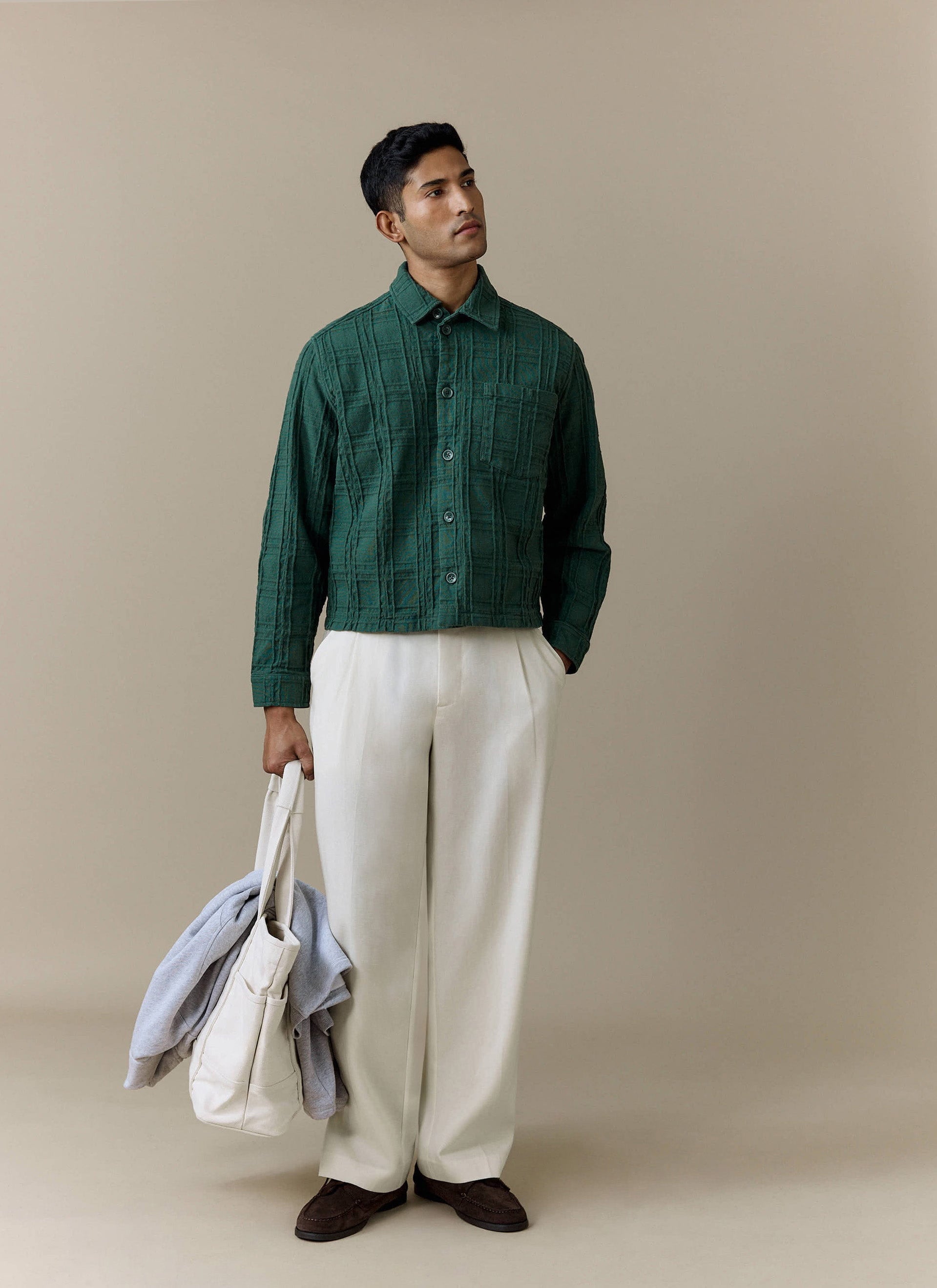 A man models the Percival Canyon Heavyweight Overshirt in Forest, styled with wide cream trousers and holding a tote bag.