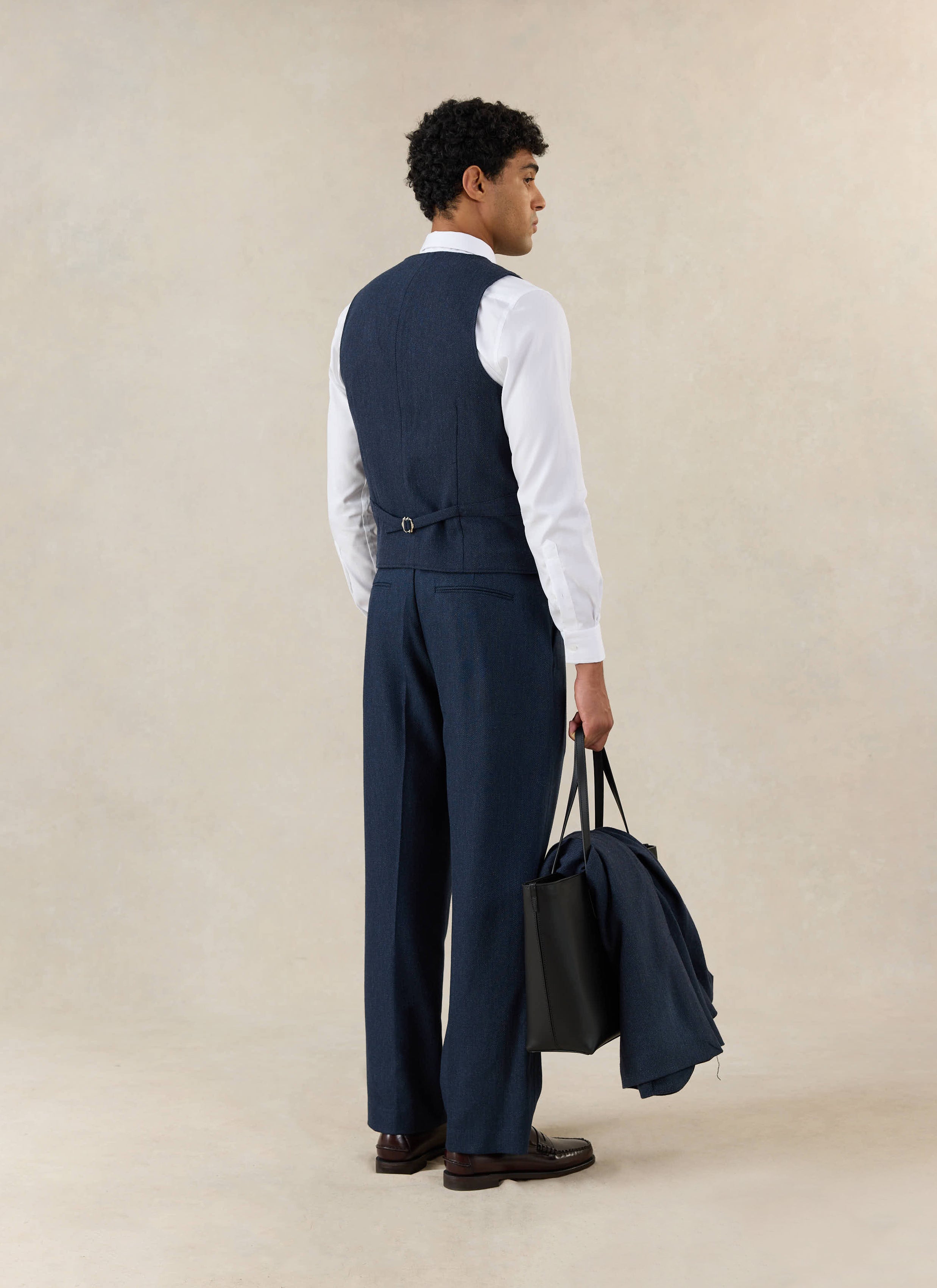 Percival Stanley Tailored Waistcoat in navy herringbone wool, shown from behind with matching trousers and a leather bag.